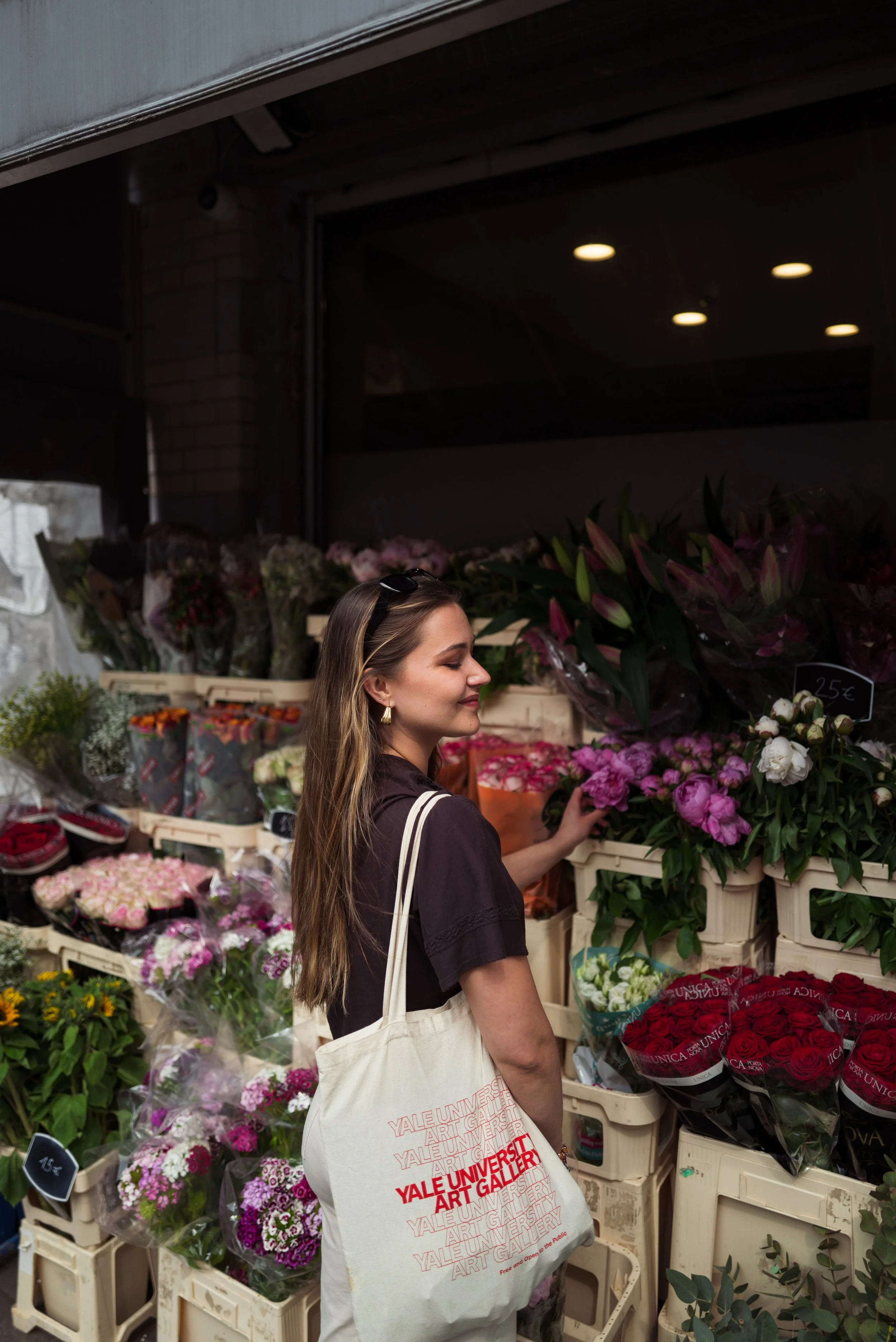 A woman shopping for flowers at an outdoor flower market, carrying a tote bag from Yale University Art Gallery.