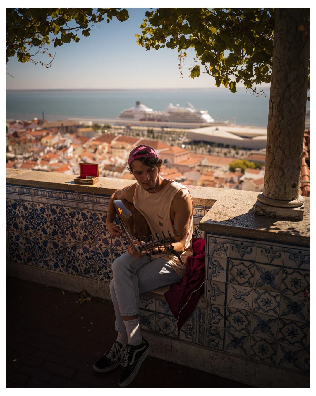 A young man sitting on a tiled ledge playing a guitar, with a view of a coastal city, a cruise ship, and the ocean in the background, under a tree with green leaves.