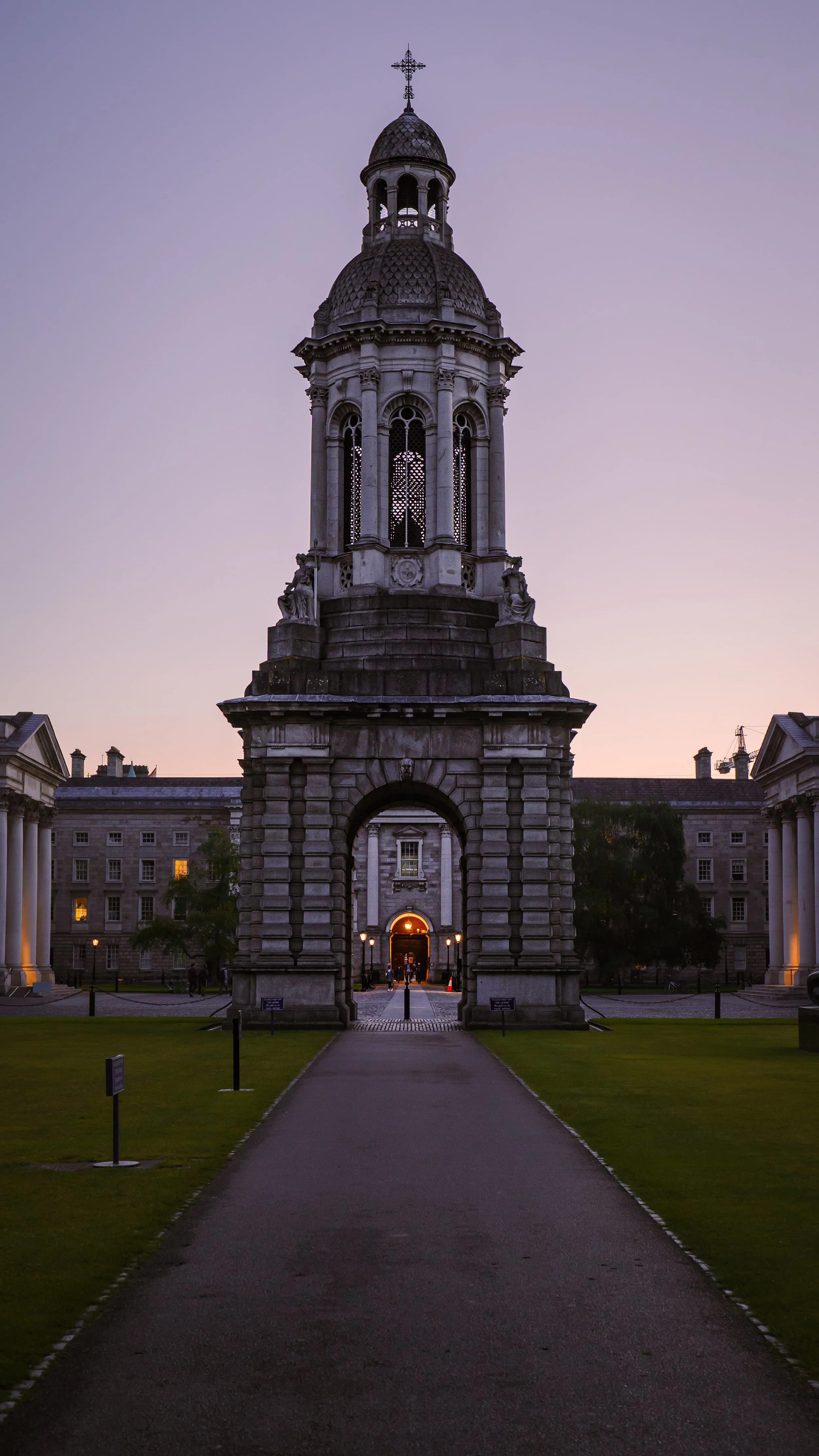 A historic stone clock tower with an arched entrance, lit lamps, and a cross on top, situated on a grassy area with pathways and classical buildings in the background at dusk.