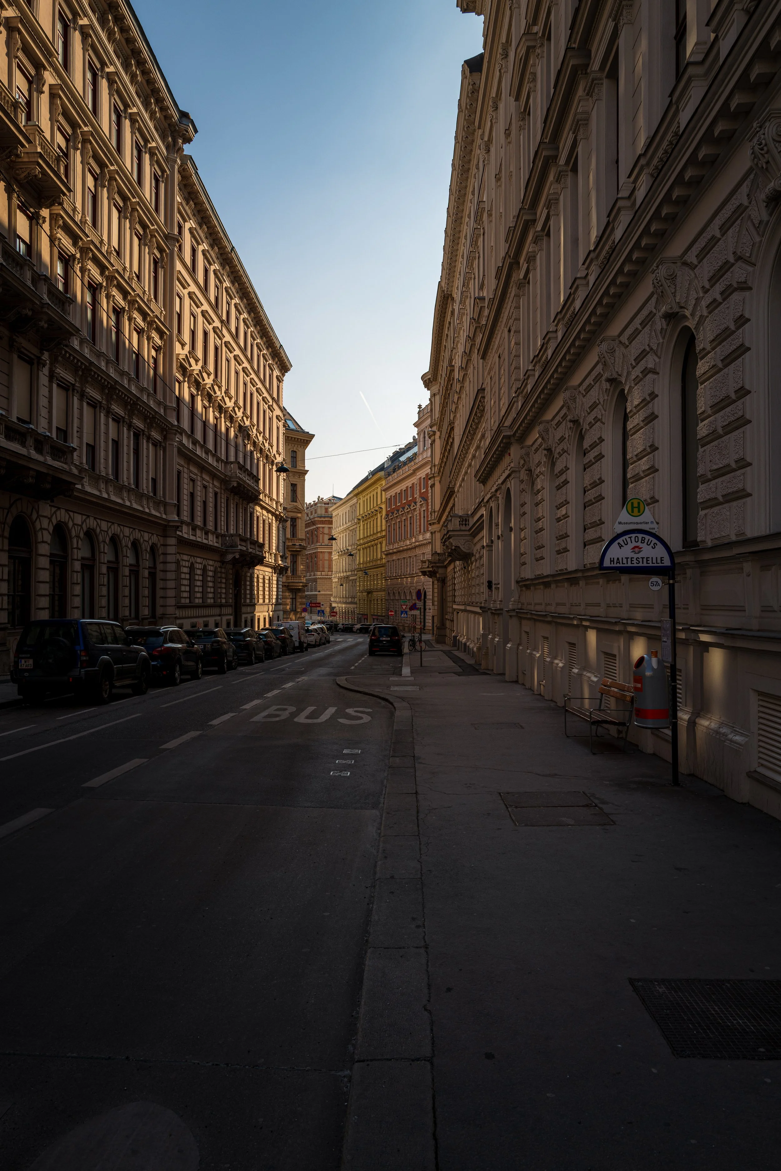A city street scene with historic buildings on both sides, parked cars lining the street, and a bus lane marked on the road. The sky is clear with a slight gradient from light to darker blue.