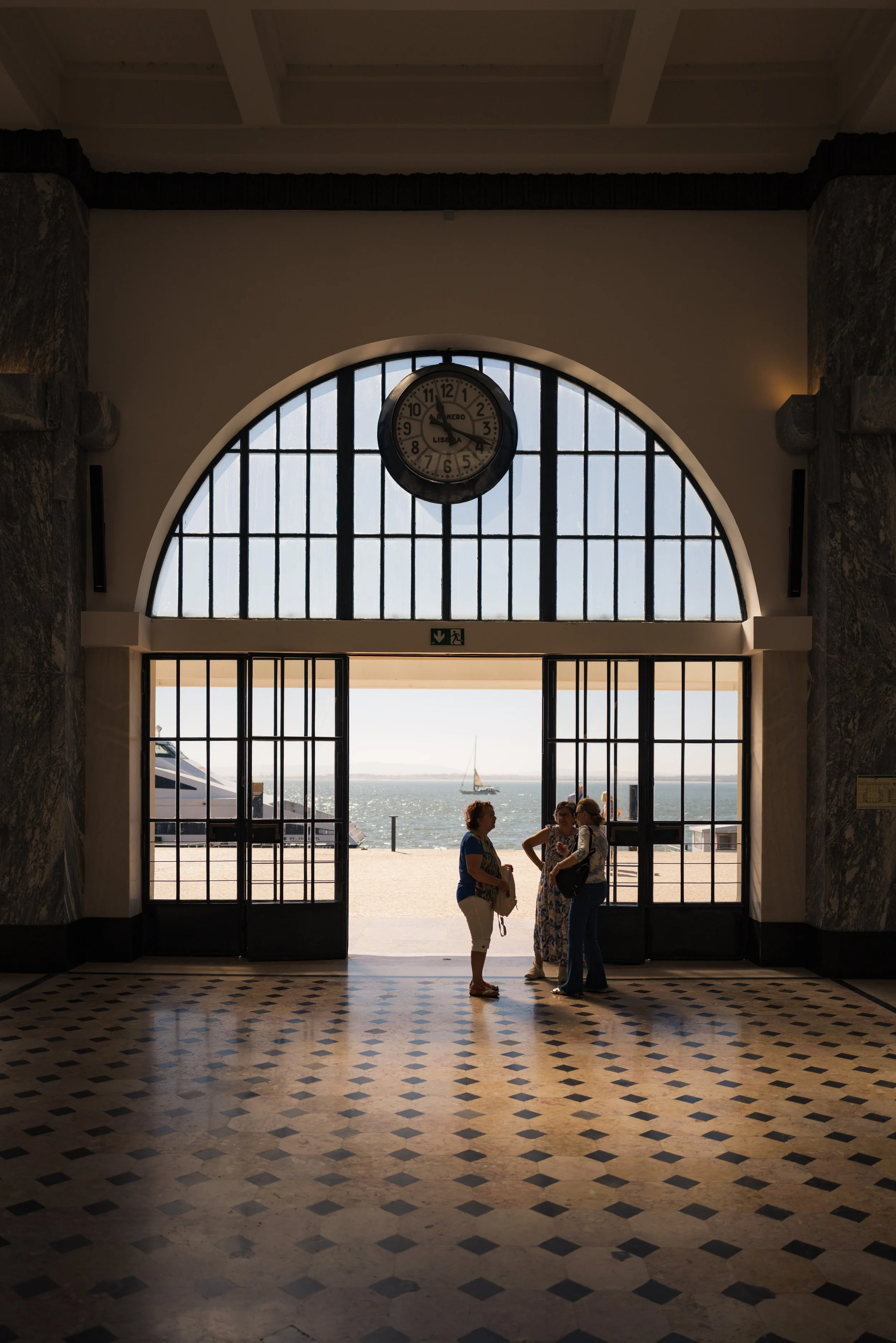 People standing inside a building near an entrance with large glass doors, overlooking a body of water with sailboats, with a clock showing around 2:12 on the wall above.