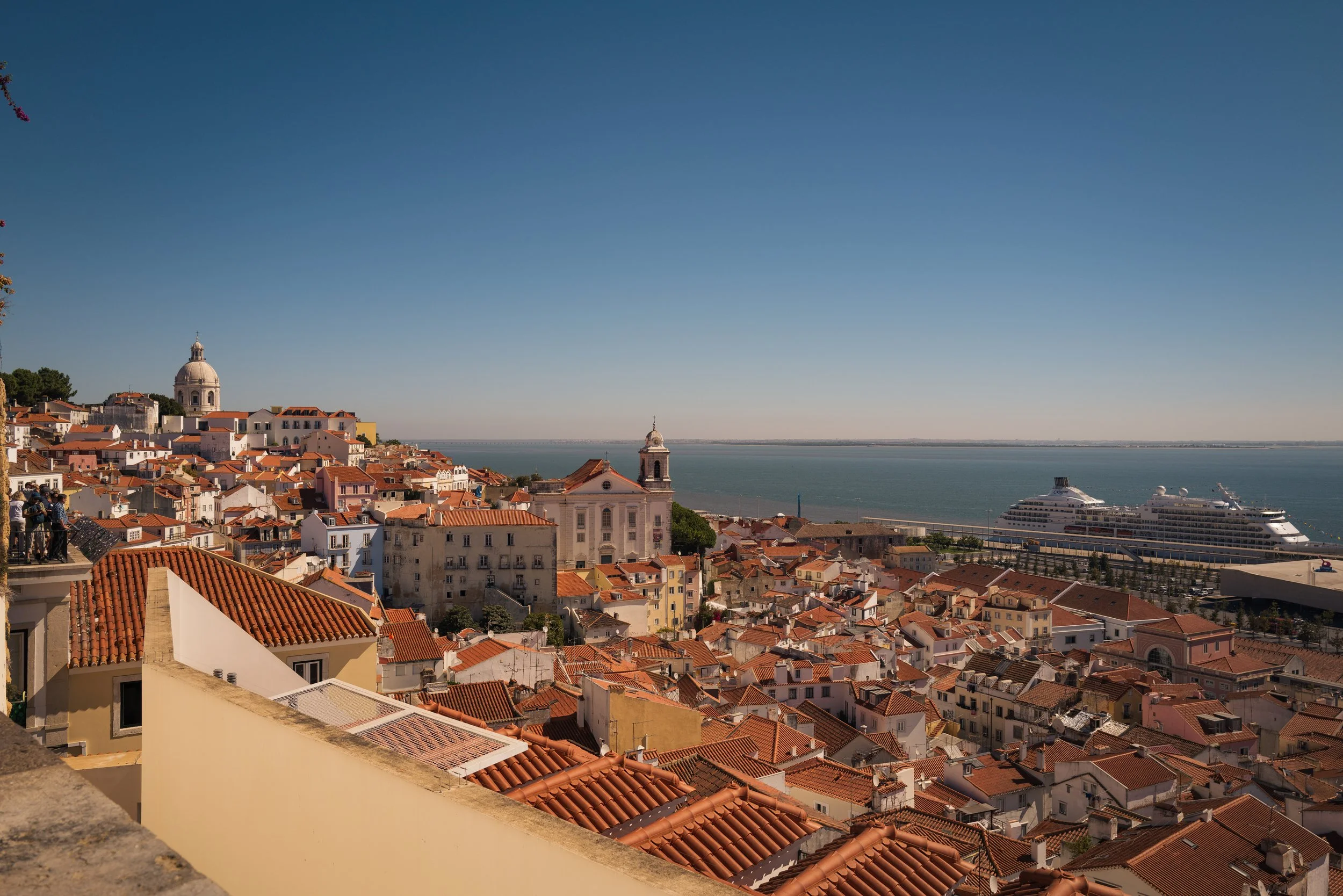 A cityscape with red-tiled rooftops, church towers, and historic buildings overlooking a large body of water with a cruise ship docked along the seaside.