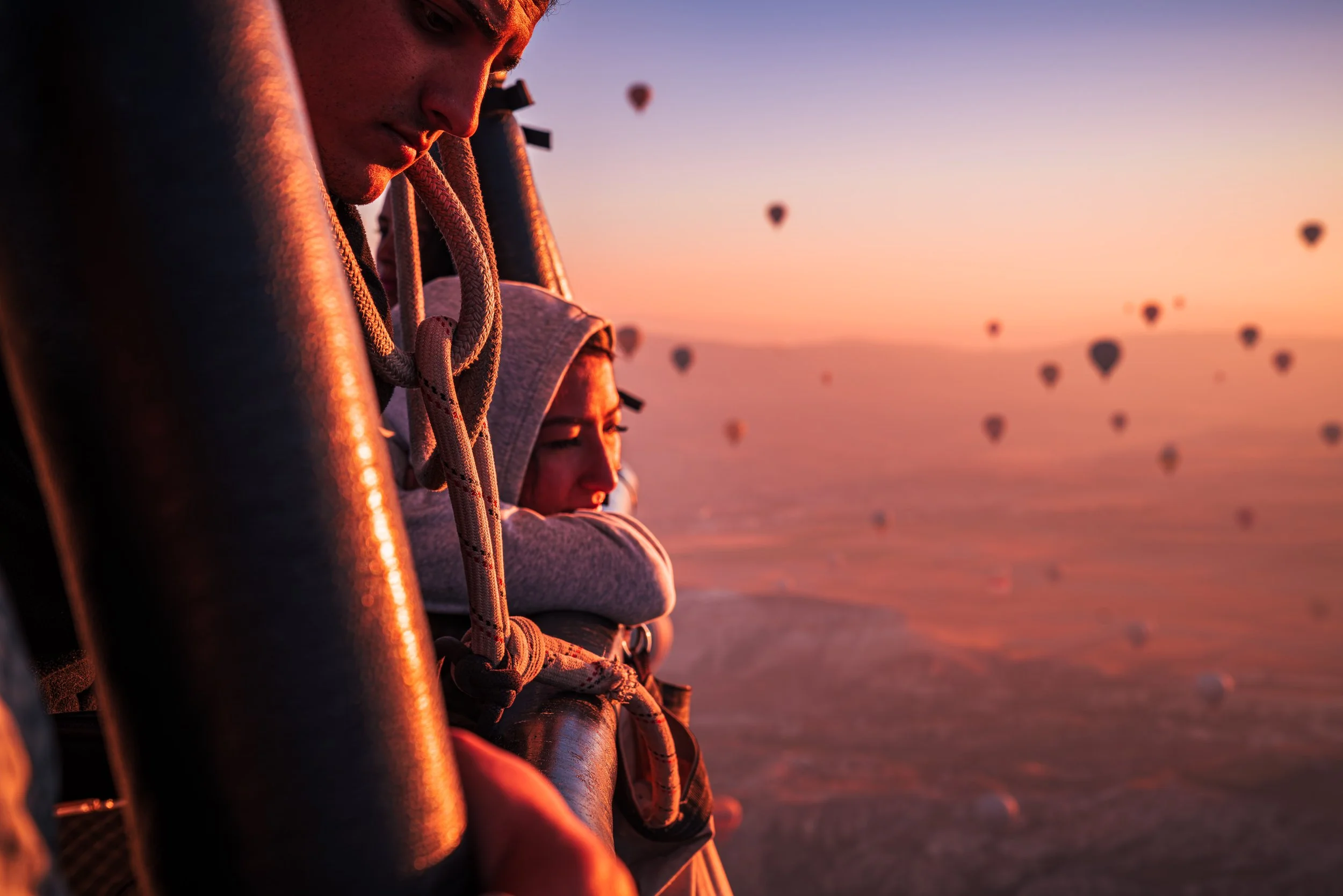 Two people in a hot air balloon at sunrise observing the landscape and hot air balloons in the sky.