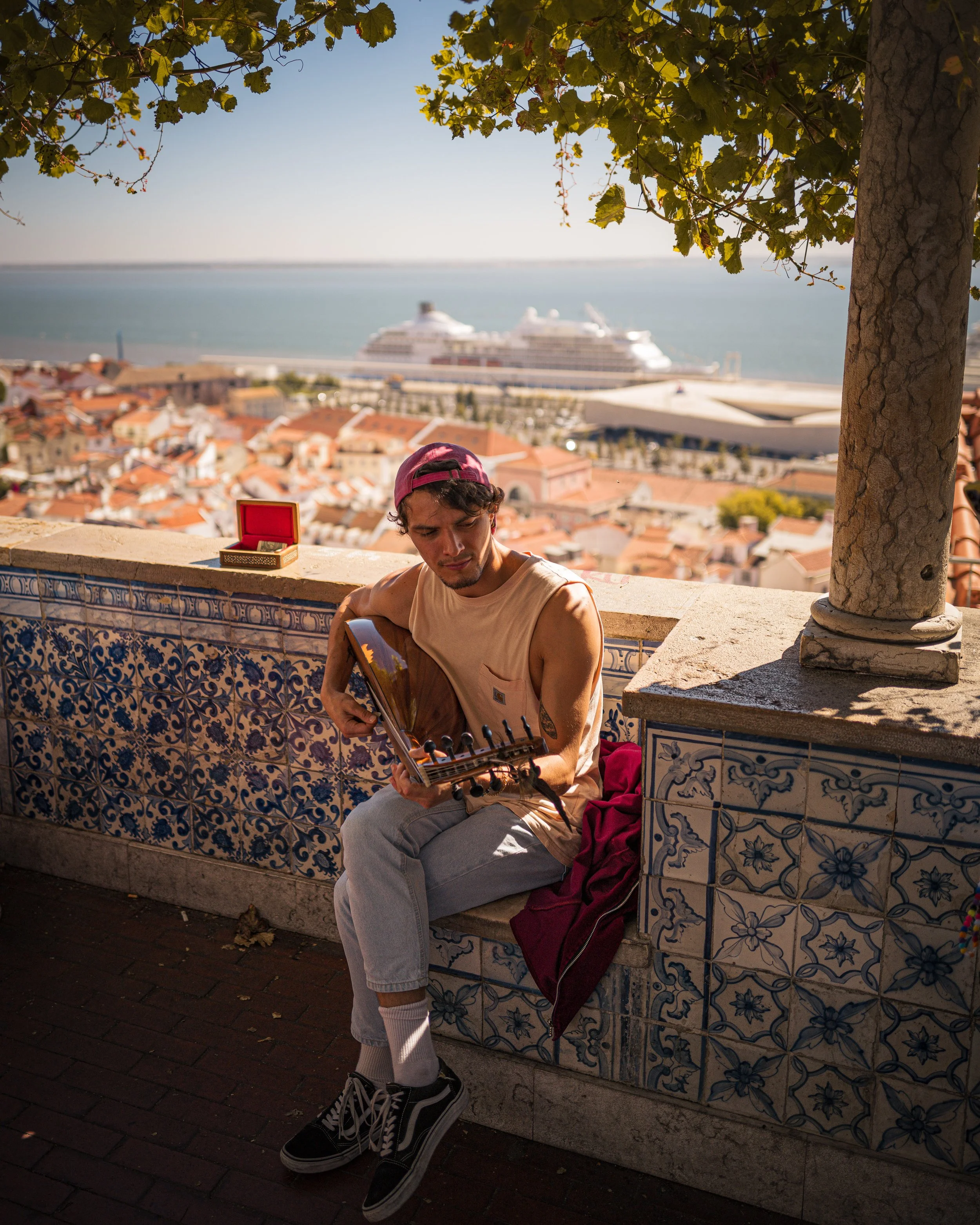 A young man playing a guitar on a terrace with decorative blue and white tiles, overlooking a cityscape with orange rooftops, a large cruise ship, and the ocean in the background, shaded by a tree.