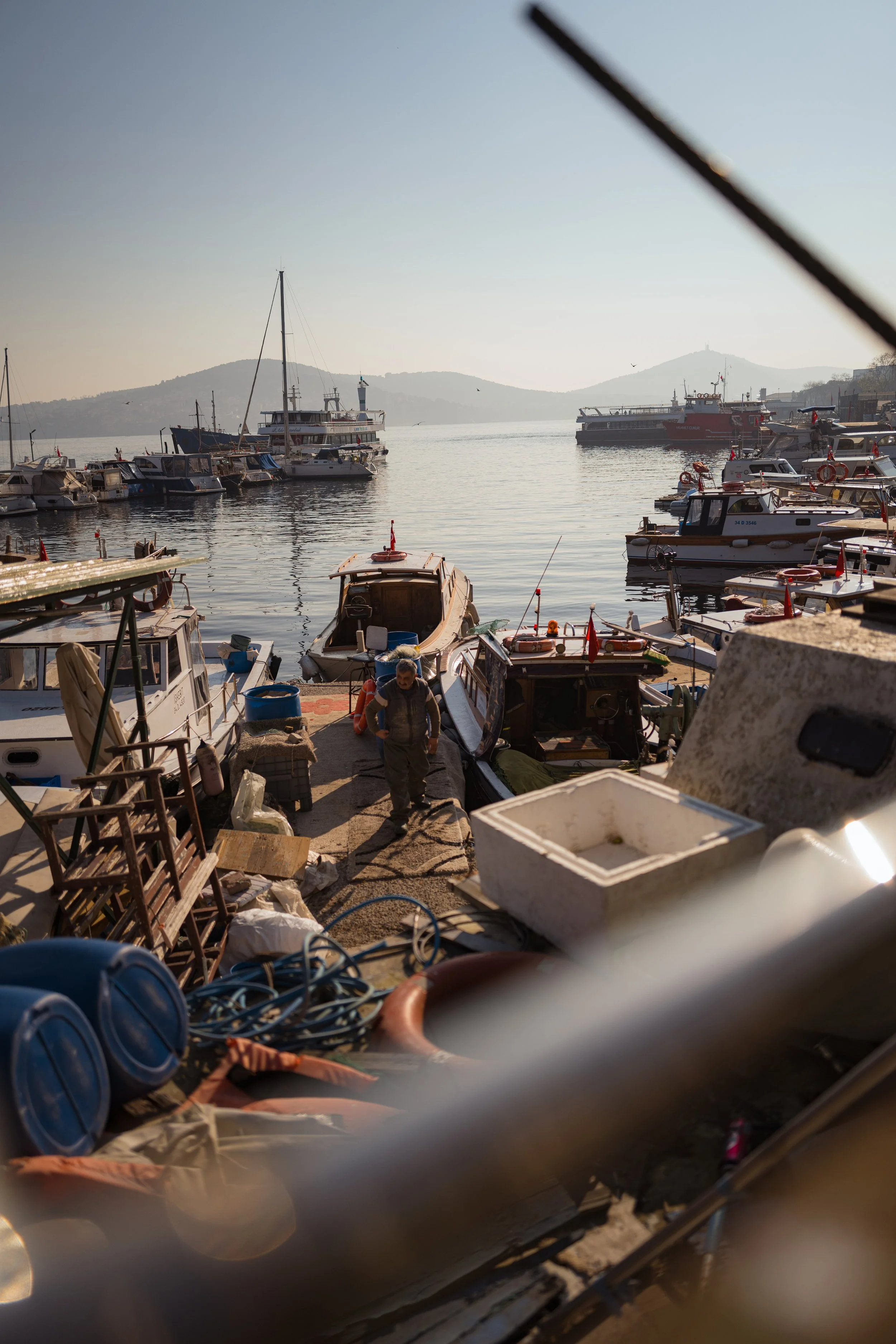 Boats docked at a marina with a person walking among the equipment, with water and hills in the background.