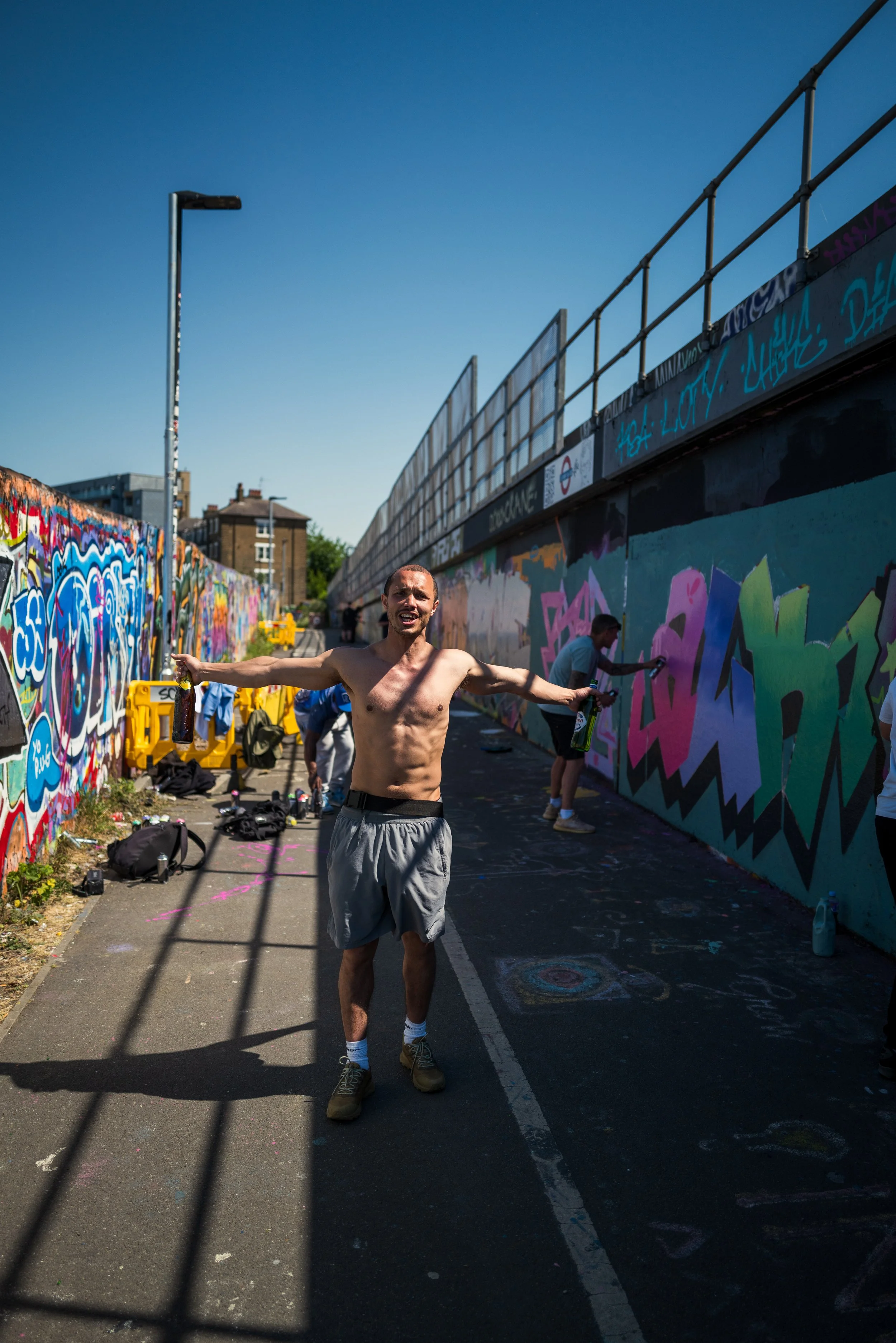 A shirtless man smiling and posing with arms outstretched in front of a graffiti-covered wall, holding bottles, while others paint graffiti on the wall behind him on a sunny day.