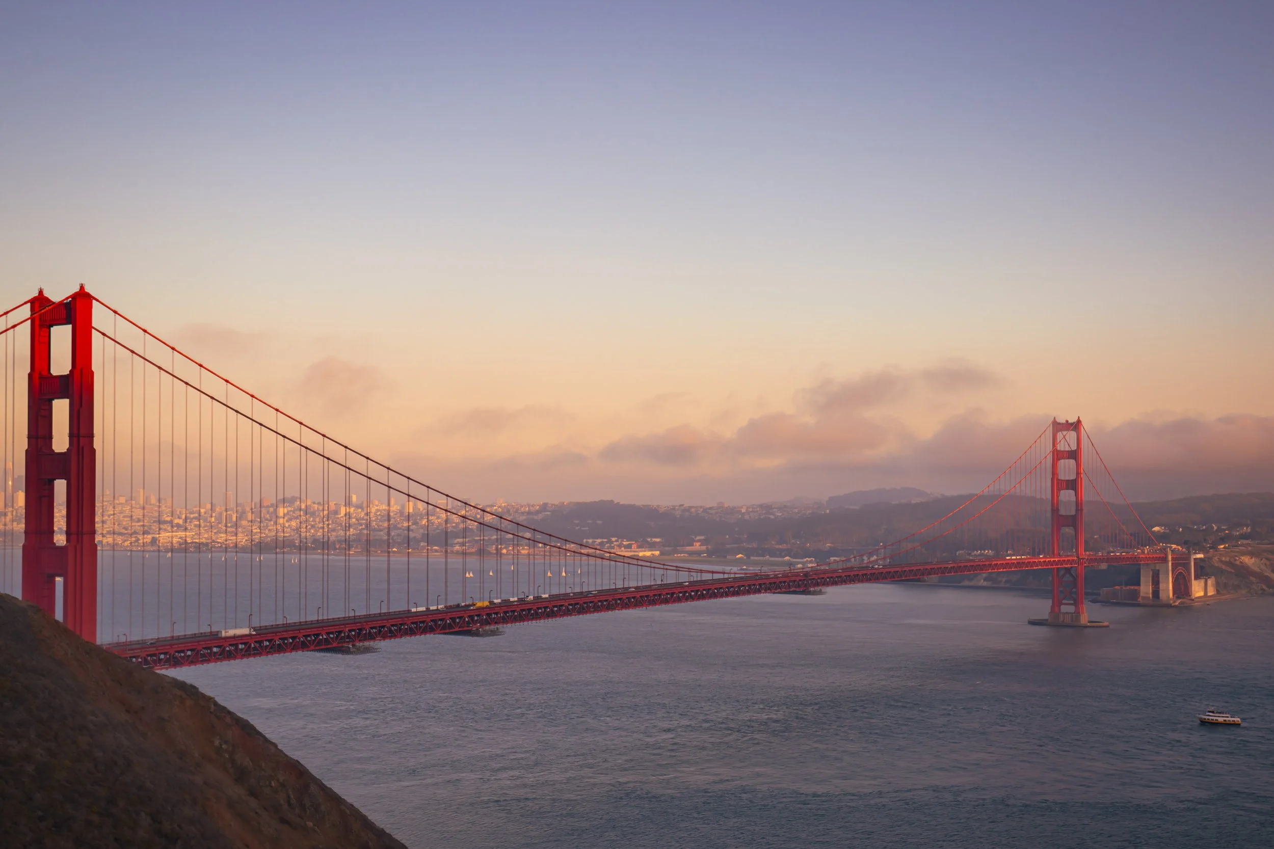 Golden Gate Bridge during sunset, with a city and hills in the background, and water with boats in the foreground.