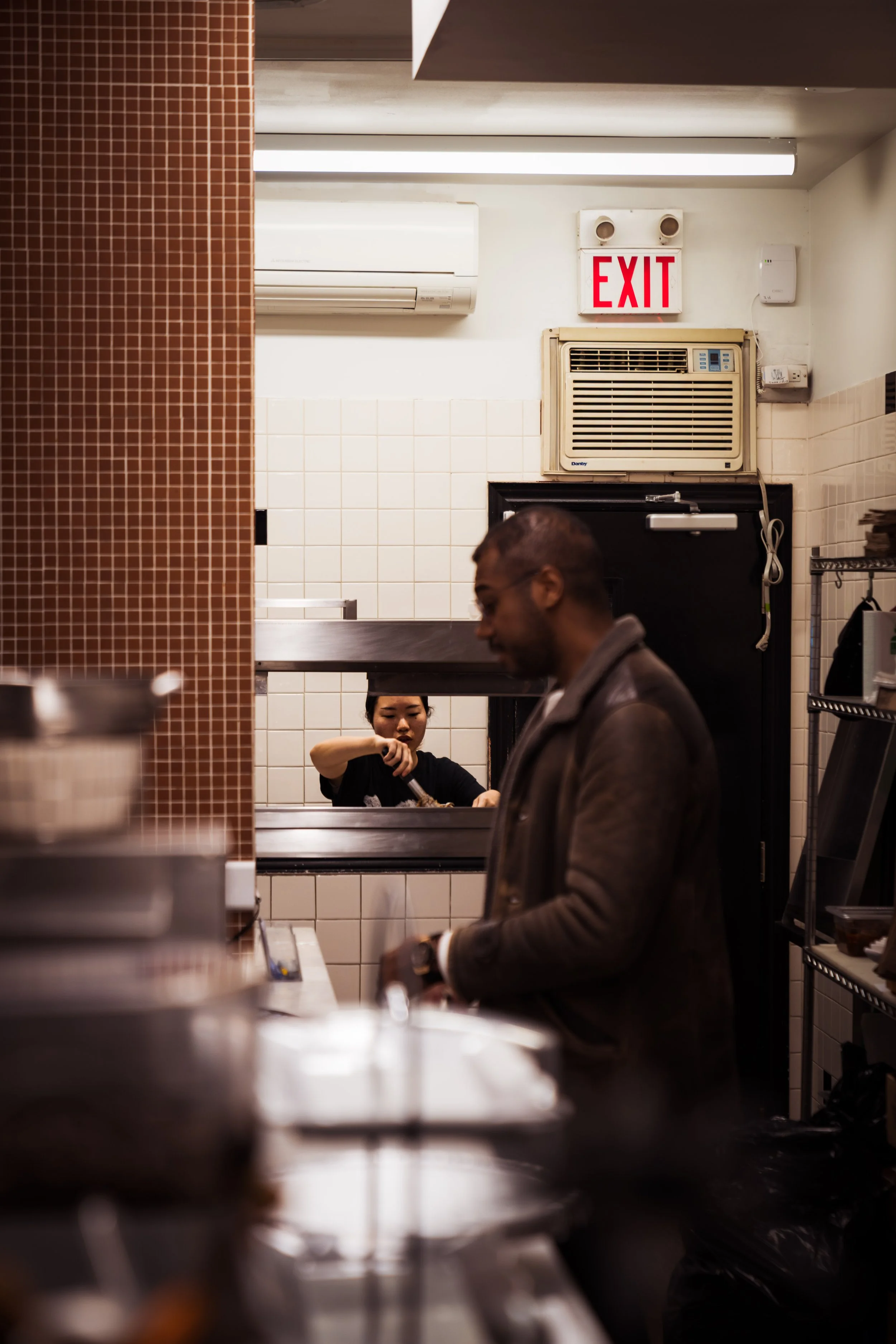 A man in a leather jacket standing in a kitchen or food service area, with a woman visible through a window serving food in the background, and a red exit sign mounted above the window.
