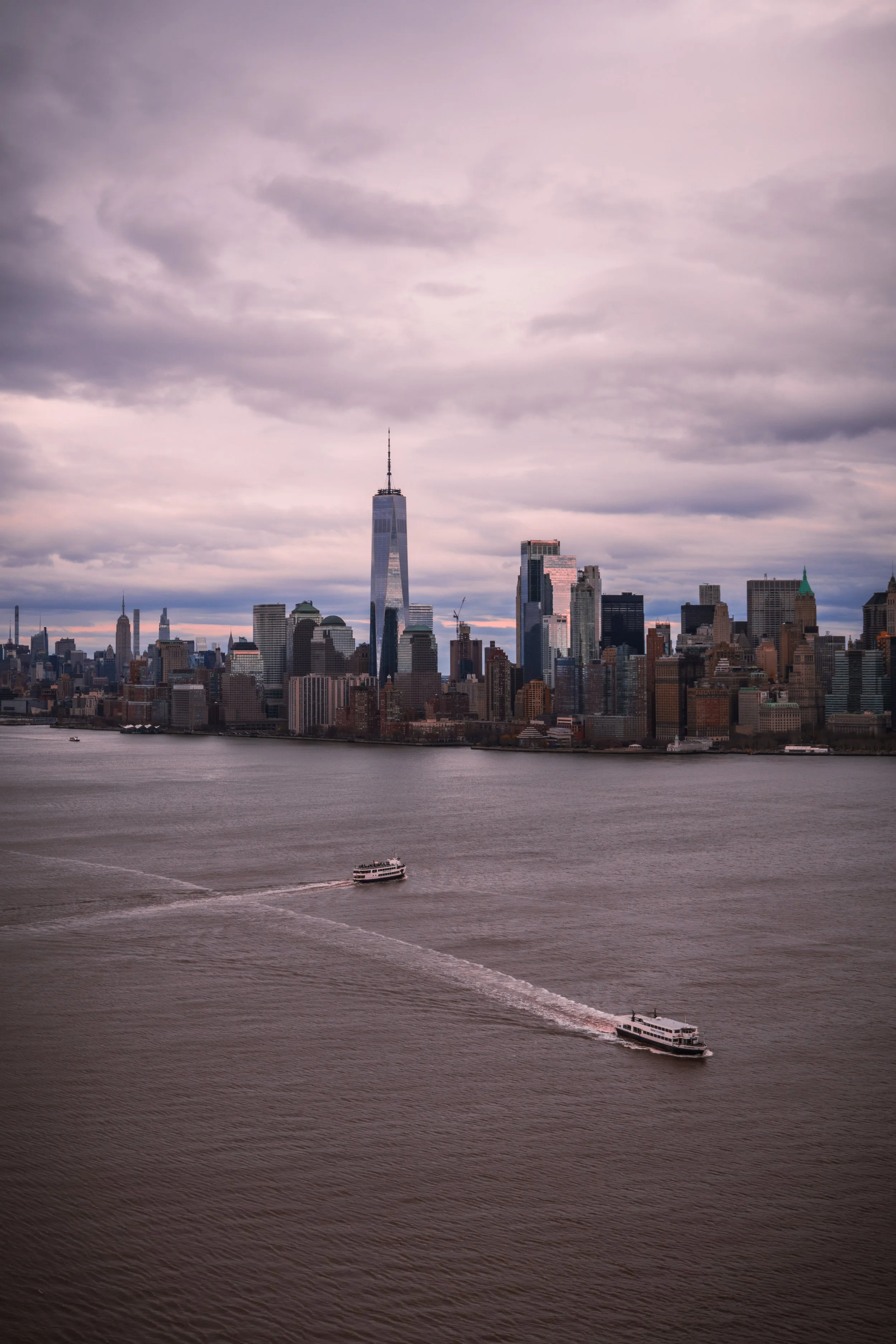 View of New York City skyline with water in foreground, featuring boats on a cloudy day.