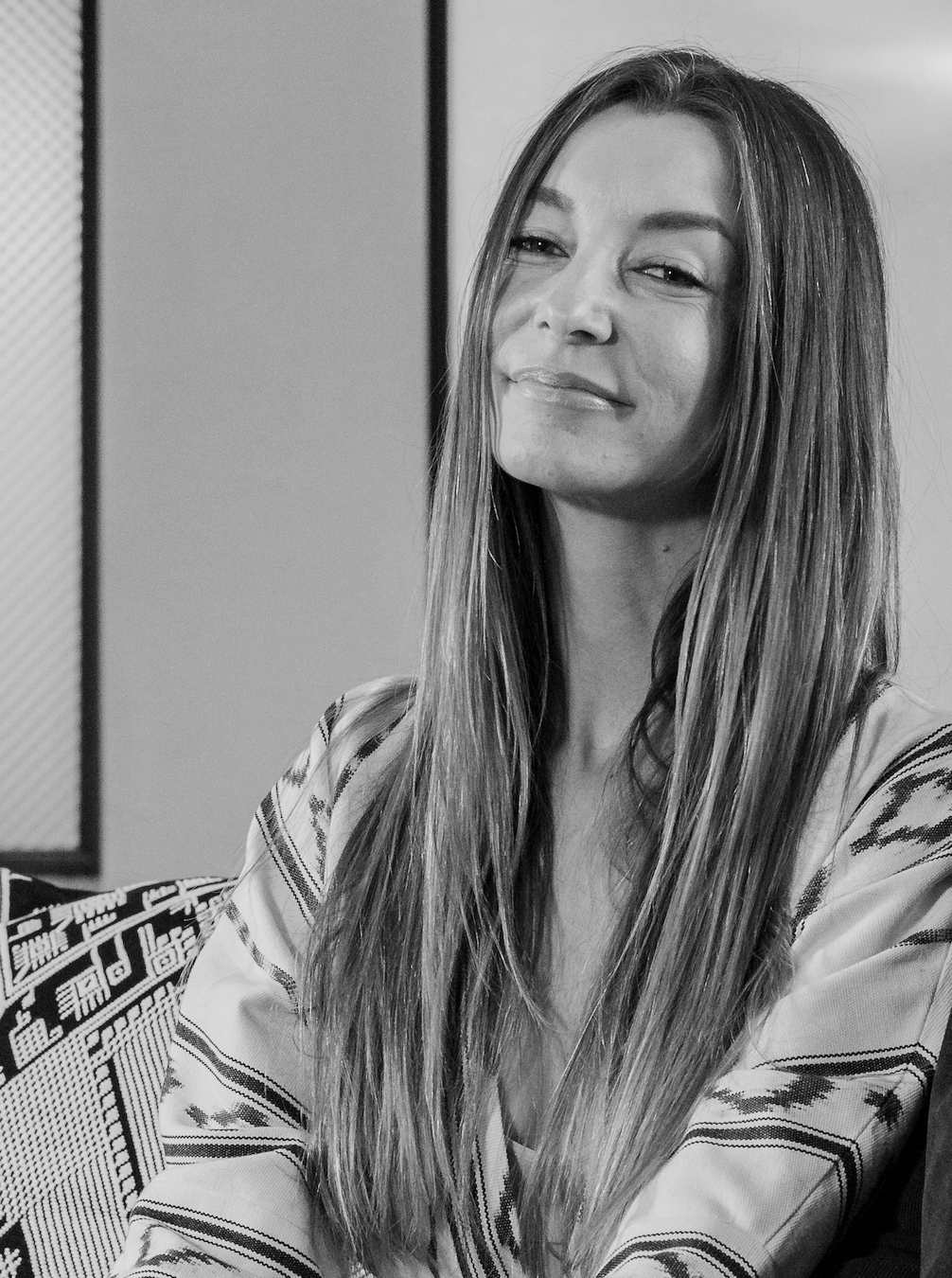 A woman with long, straight hair sitting and smiling in a modern indoor setting.