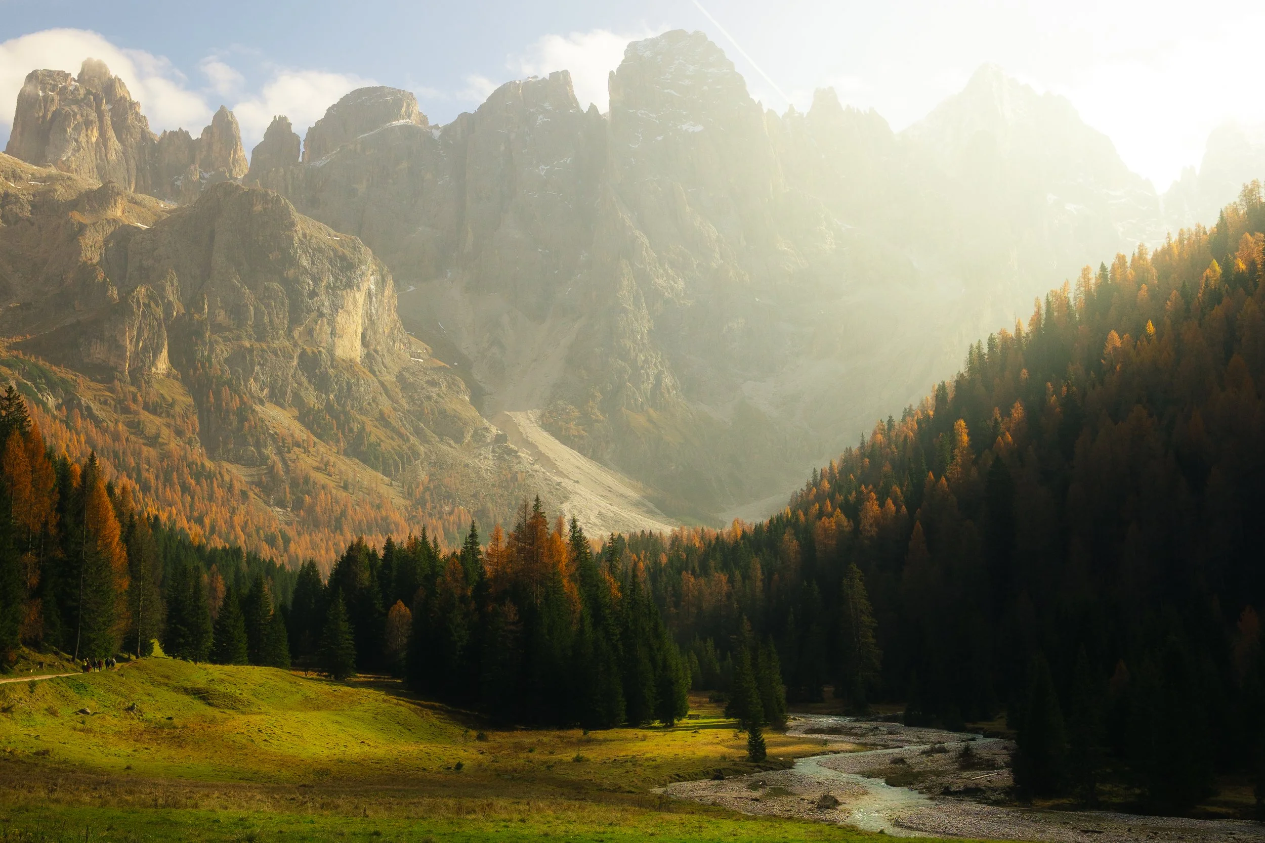 A scenic landscape of mountains with rocky peaks and rounder hills, covered in trees with autumn colors, a green meadow in the foreground, and a river running through it.