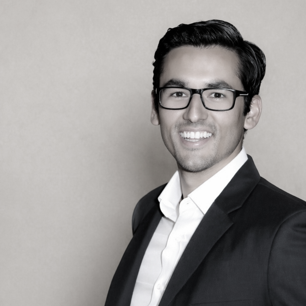 A professional headshot of a man with dark hair, wearing glasses, a white shirt, and a black blazer, smiling against a plain background.