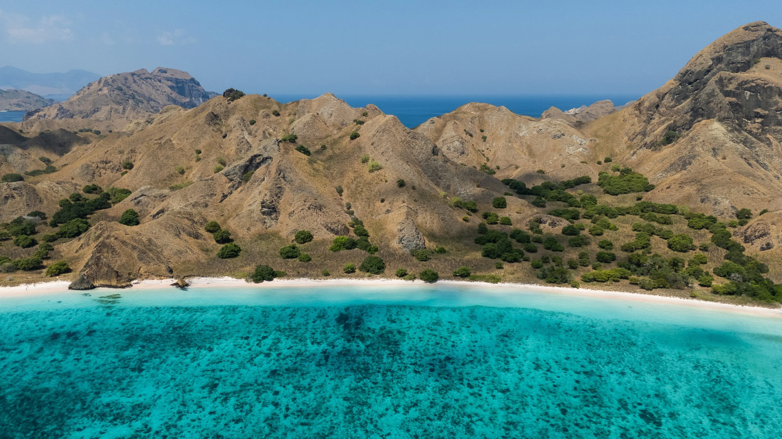 A landscape of coastal mountains with dry grass and scattered green bushes, a white sandy beach, and clear turquoise ocean water.
