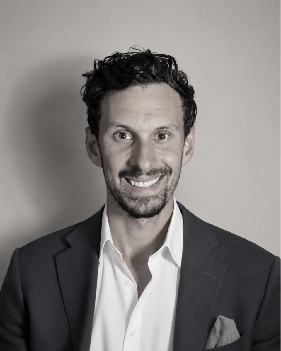 A smiling man with dark, curly hair, wearing a dark suit jacket and white shirt, against a plain gray background.