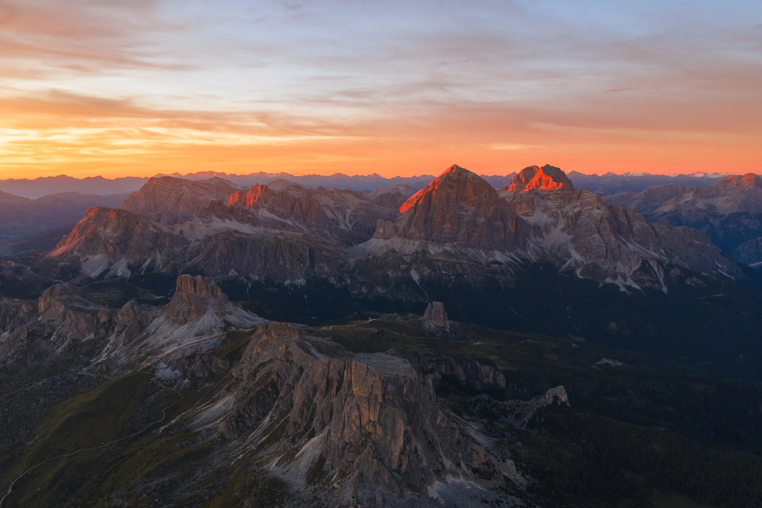 A scenic view of mountain peaks during sunset with orange and blue sky, rugged terrain, and valley pathways.
