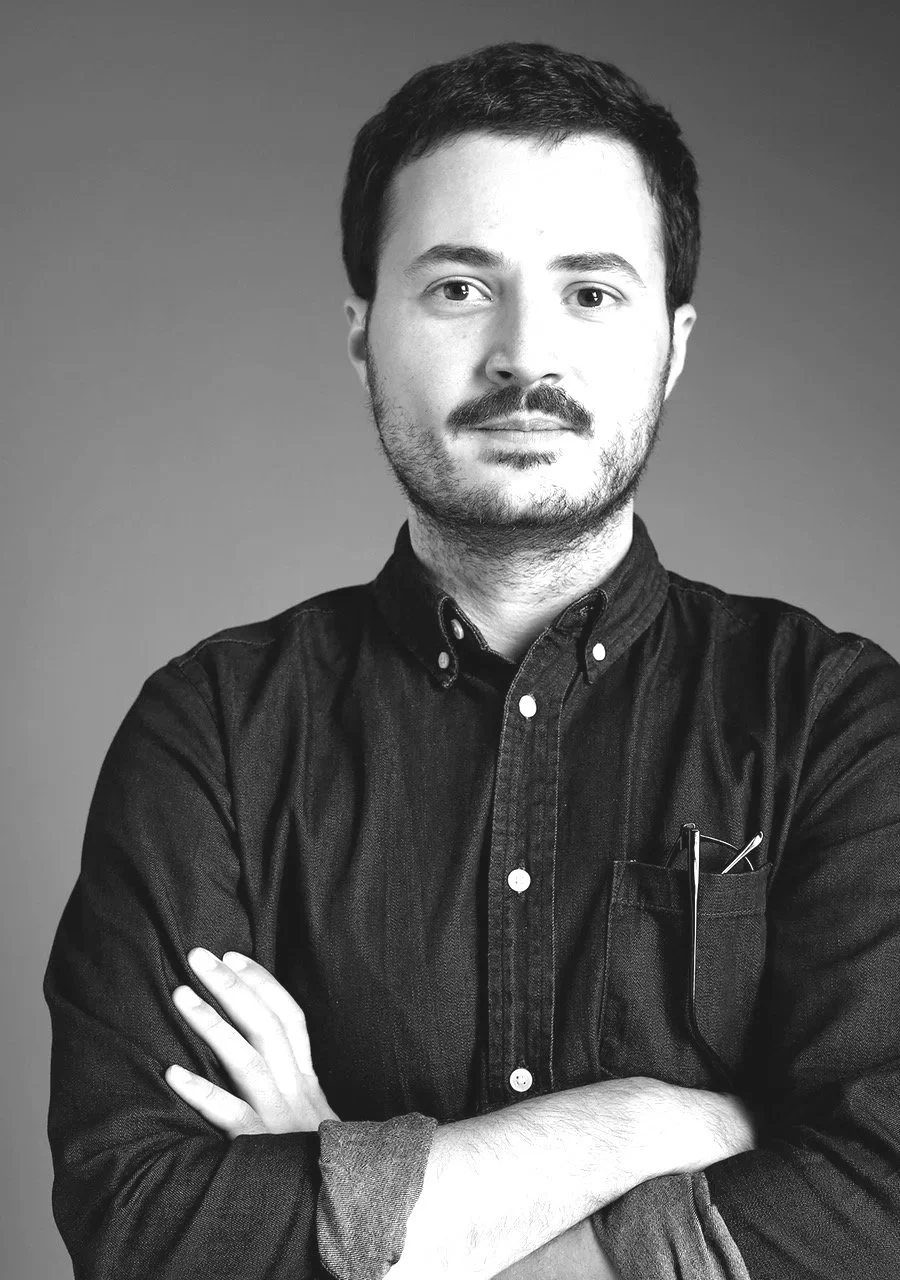 Black and white portrait of a man with short dark hair, facial hair, wearing a dark button-up shirt with rolled-up sleeves, standing with arms crossed against a plain background.