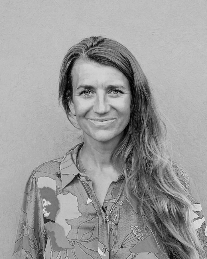 Black and white portrait of a smiling woman with long, wavy hair wearing a patterned shirt, standing against a plain wall.