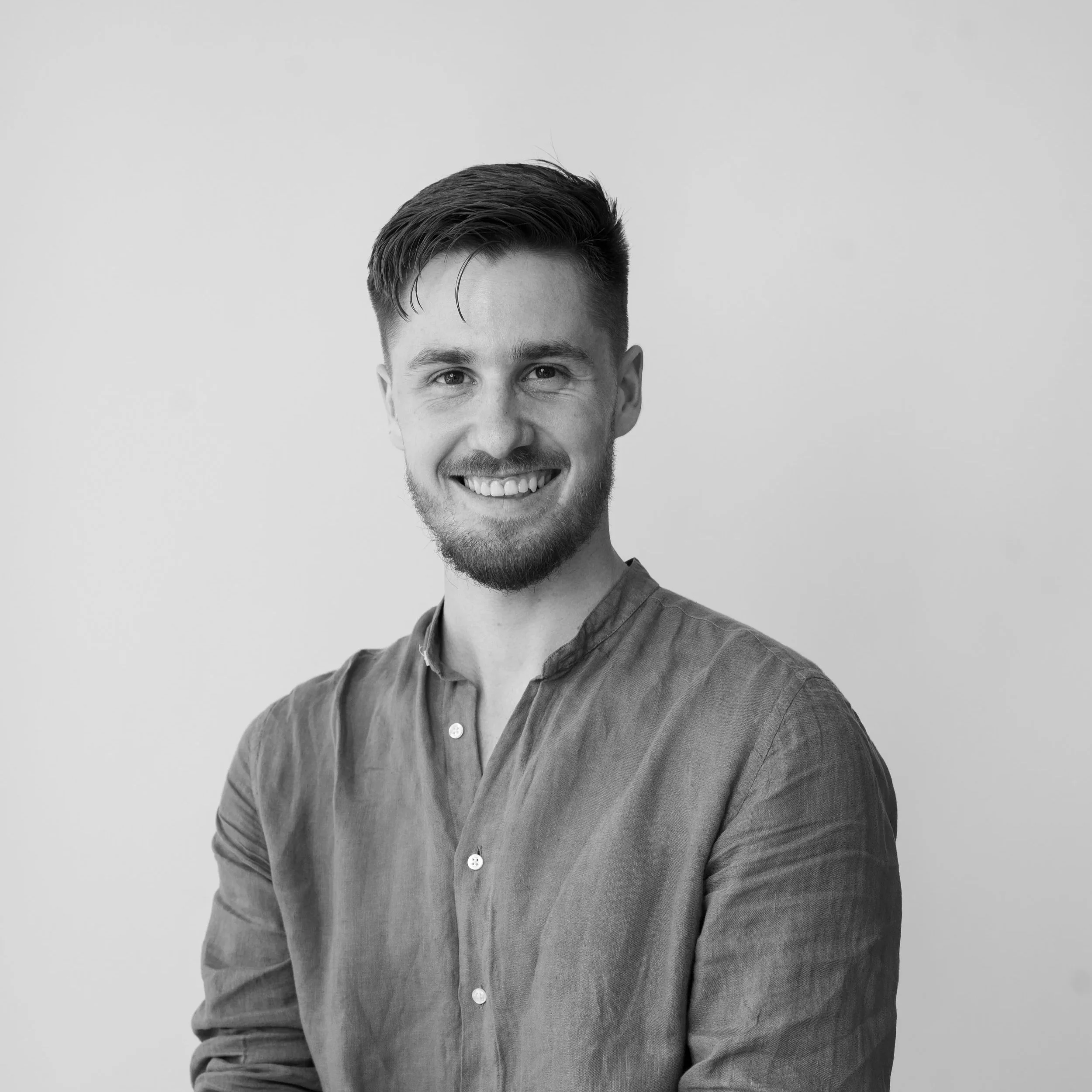 A black-and-white photo of a man smiling, with short dark hair, a beard, in a button-up shirt, standing against a plain light gray background.