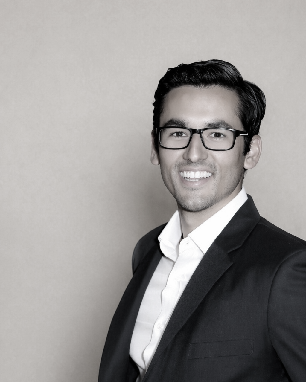 A smiling man with dark hair, glasses, wearing a suit and white shirt, standing against a plain wall.