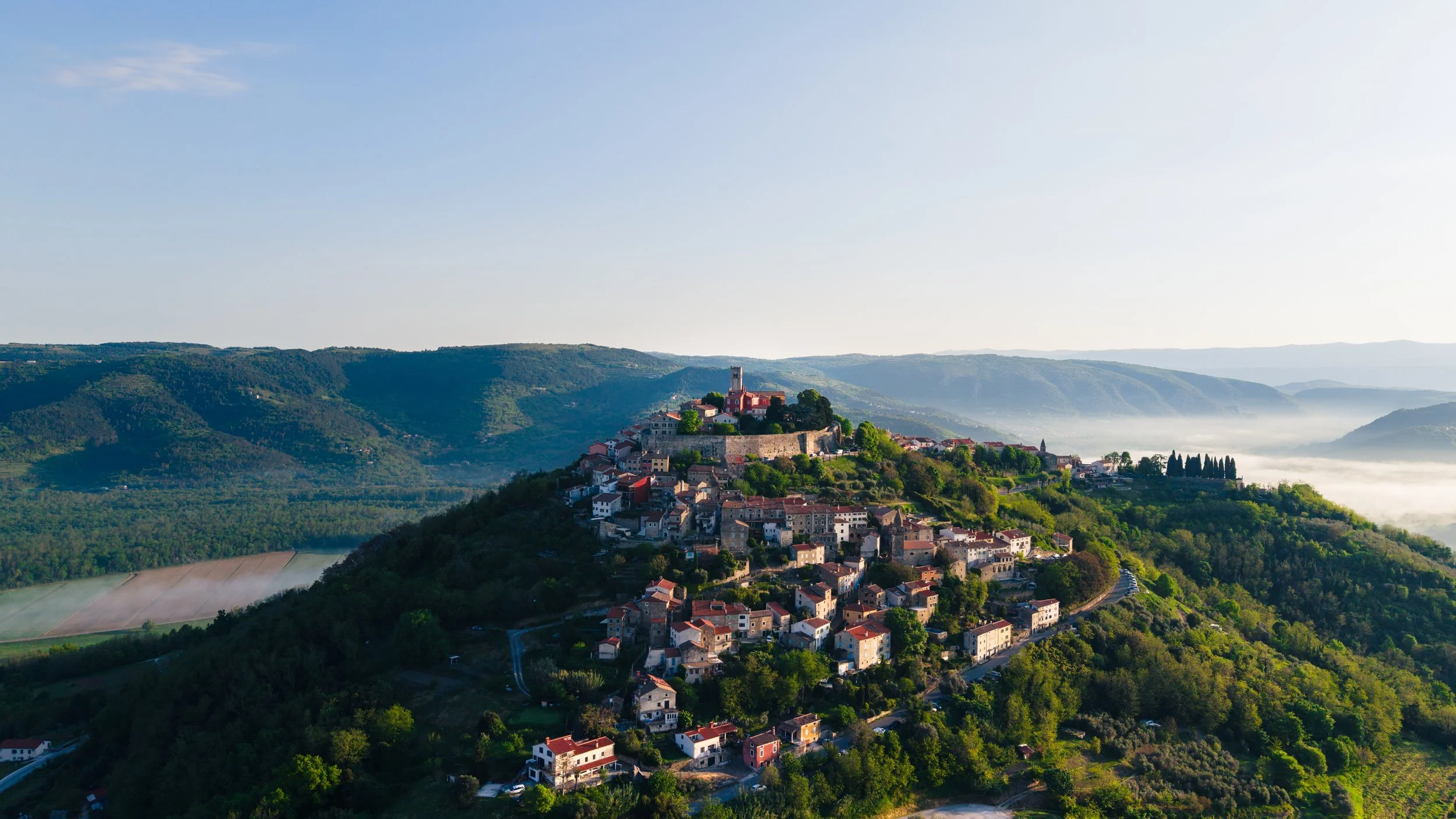 A scenic aerial view of a hillside village with a castle on top, surrounded by green hills and valleys under a clear sky with some clouds.