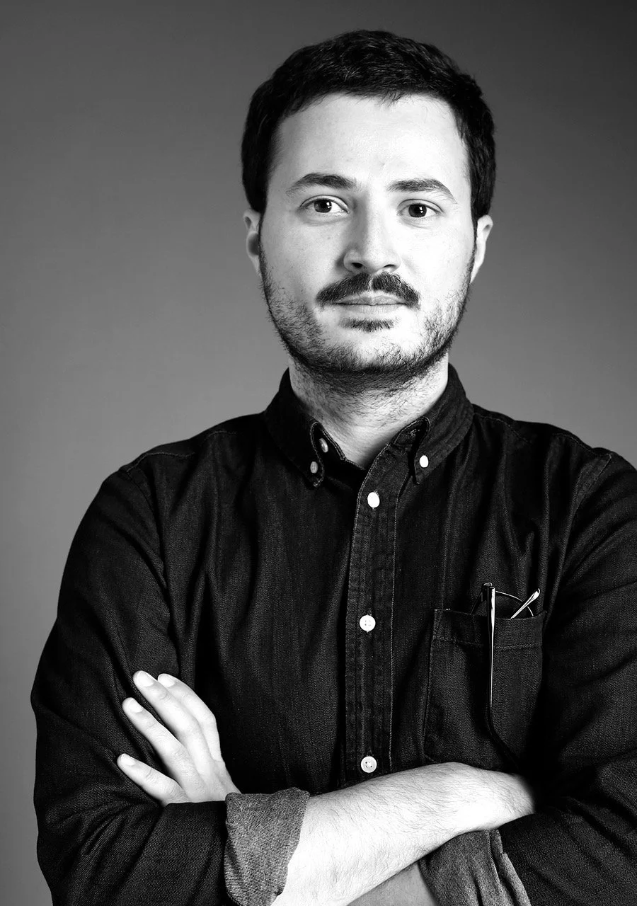 Black and white portrait of a man with short dark hair, facial hair, wearing a black button-up shirt with rolled-up sleeves, standing with arms crossed against a gradient background.