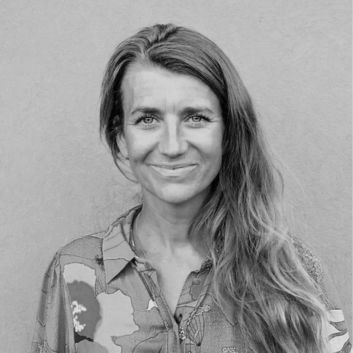 A woman with long, wavy hair and light-colored eyes, smiling, wearing a patterned shirt, standing against a plain wall.