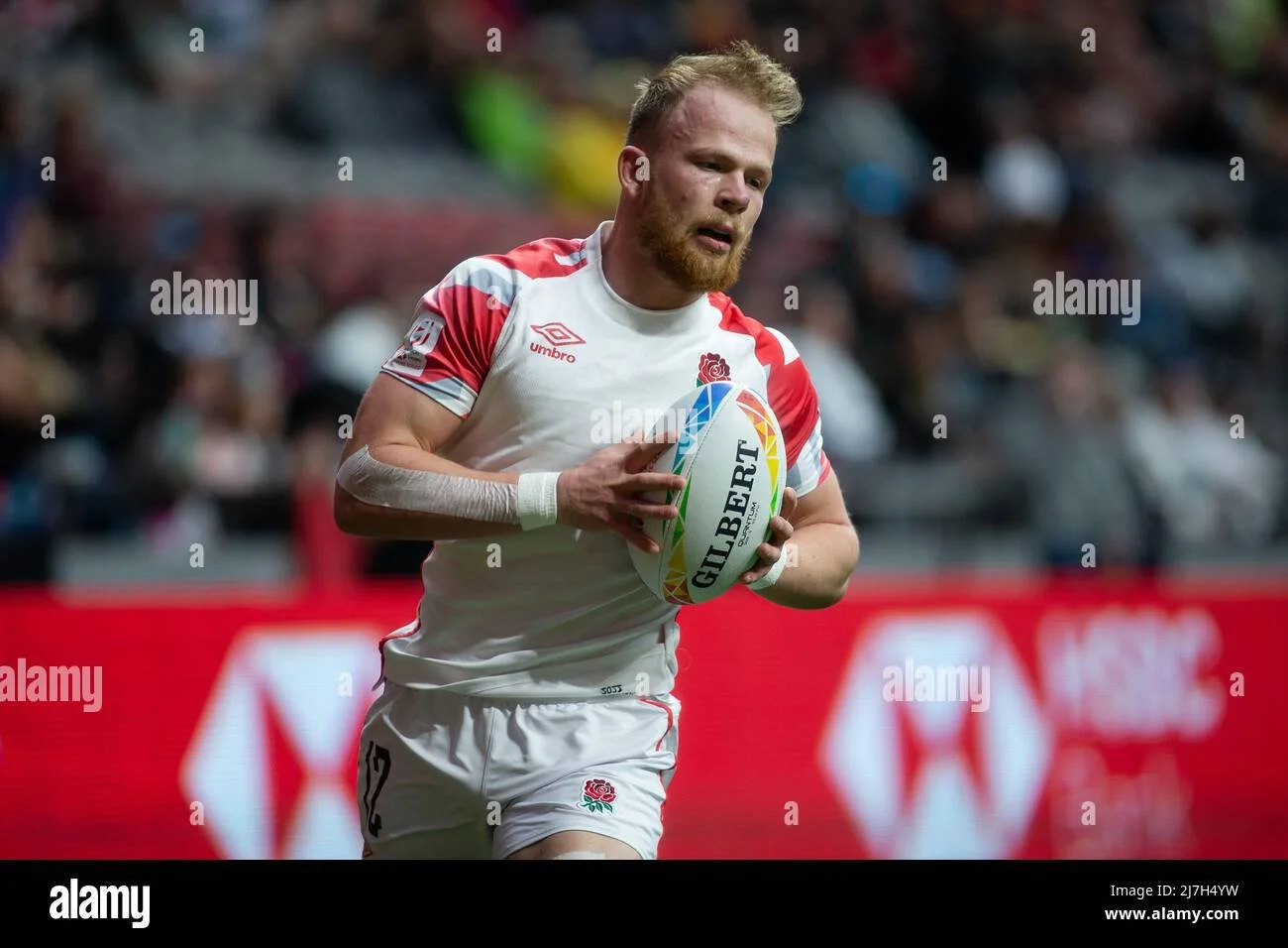 A rugby player in a white and red uniform holding a Gilbert rugby ball on a field with a crowd in the background.