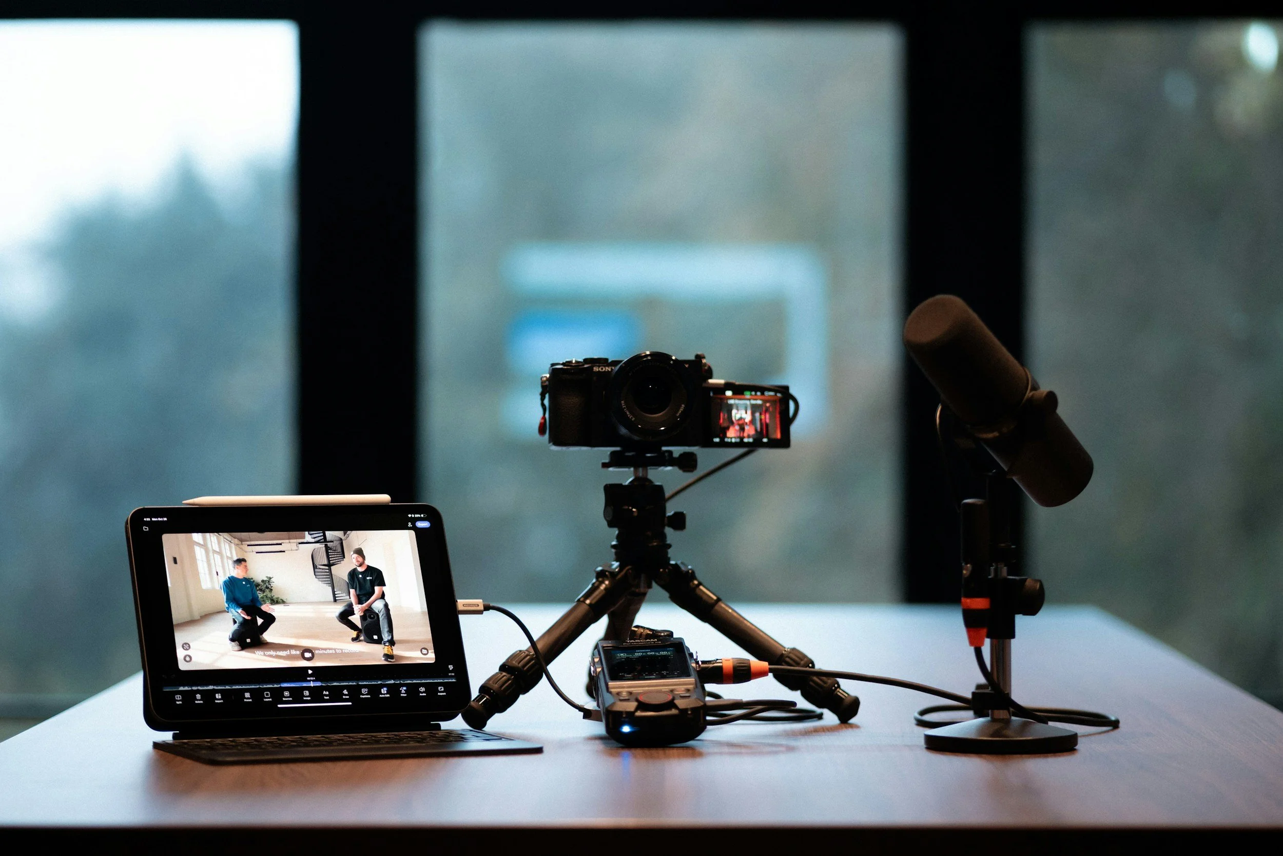 Video recording setup with a tablet showing two people sitting and talking, a camera on a tripod, a microphone, and audio equipment on a wooden table in front of large windows.