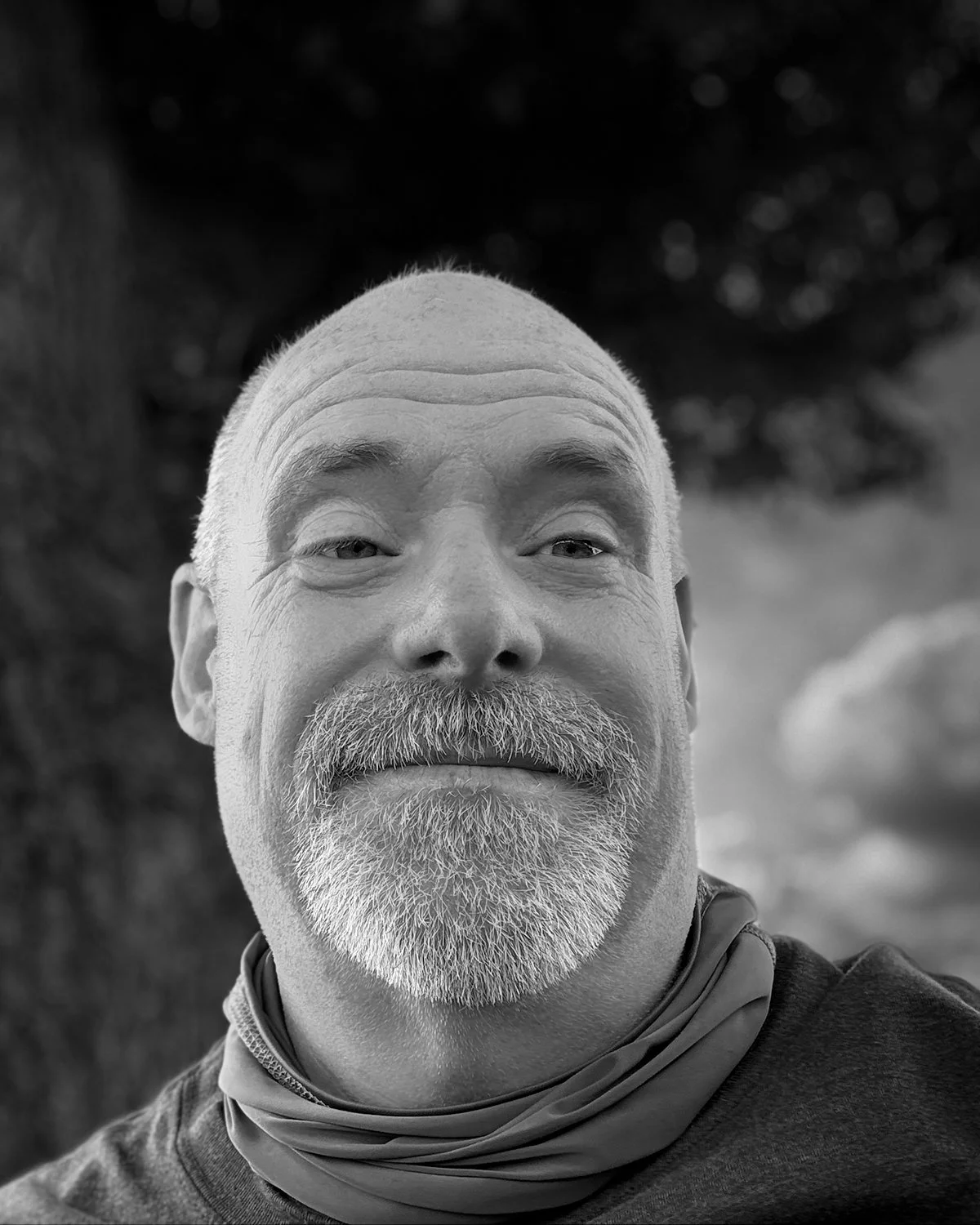A black and white close-up photo of a smiling middle-aged man with a beard and short hair, outdoors with blurred trees in the background.