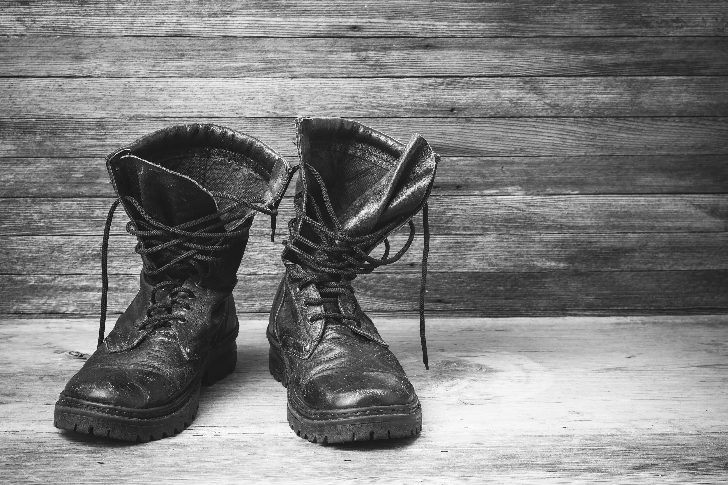 Black leather work boots with scuffed toes on a wooden surface with a wooden plank wall in the background.