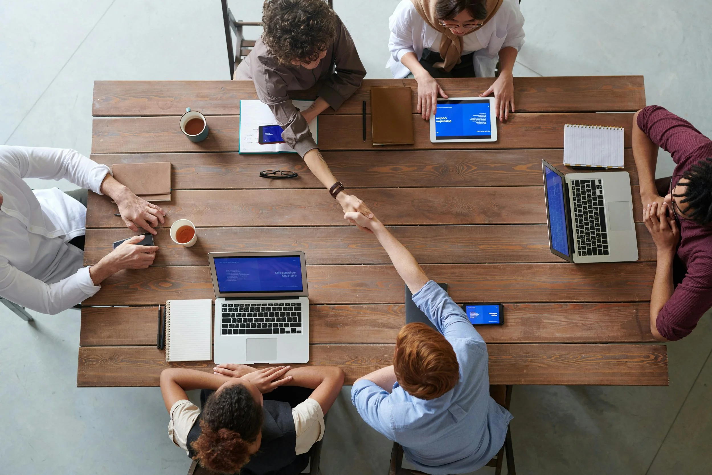 Six people around a wooden table in a meeting, shaking hands, with laptops, notebooks, and cups of coffee.