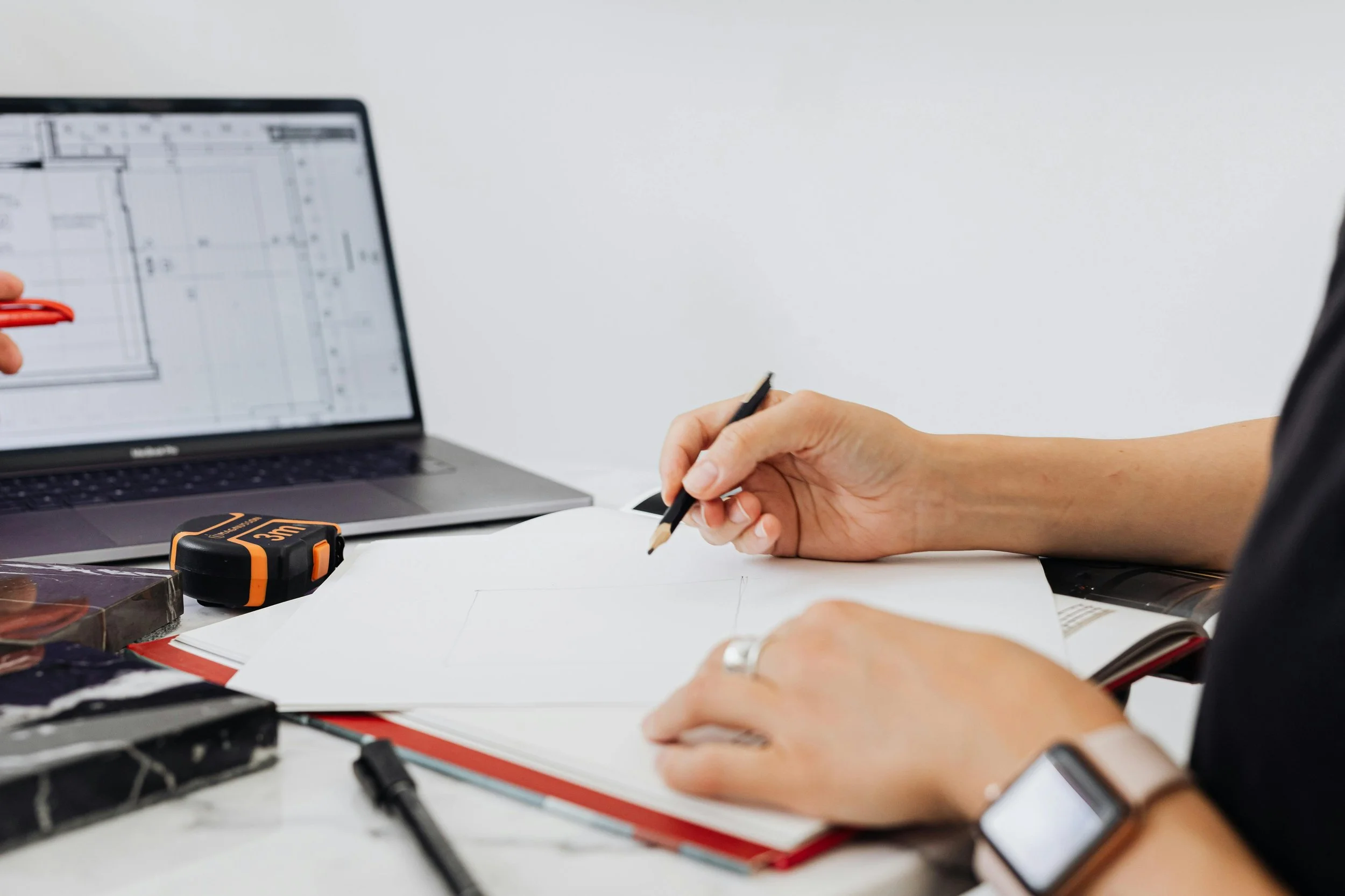 A person working at a desk with a laptop, notebook, and drawing tools, including a pencil and a tape measure.