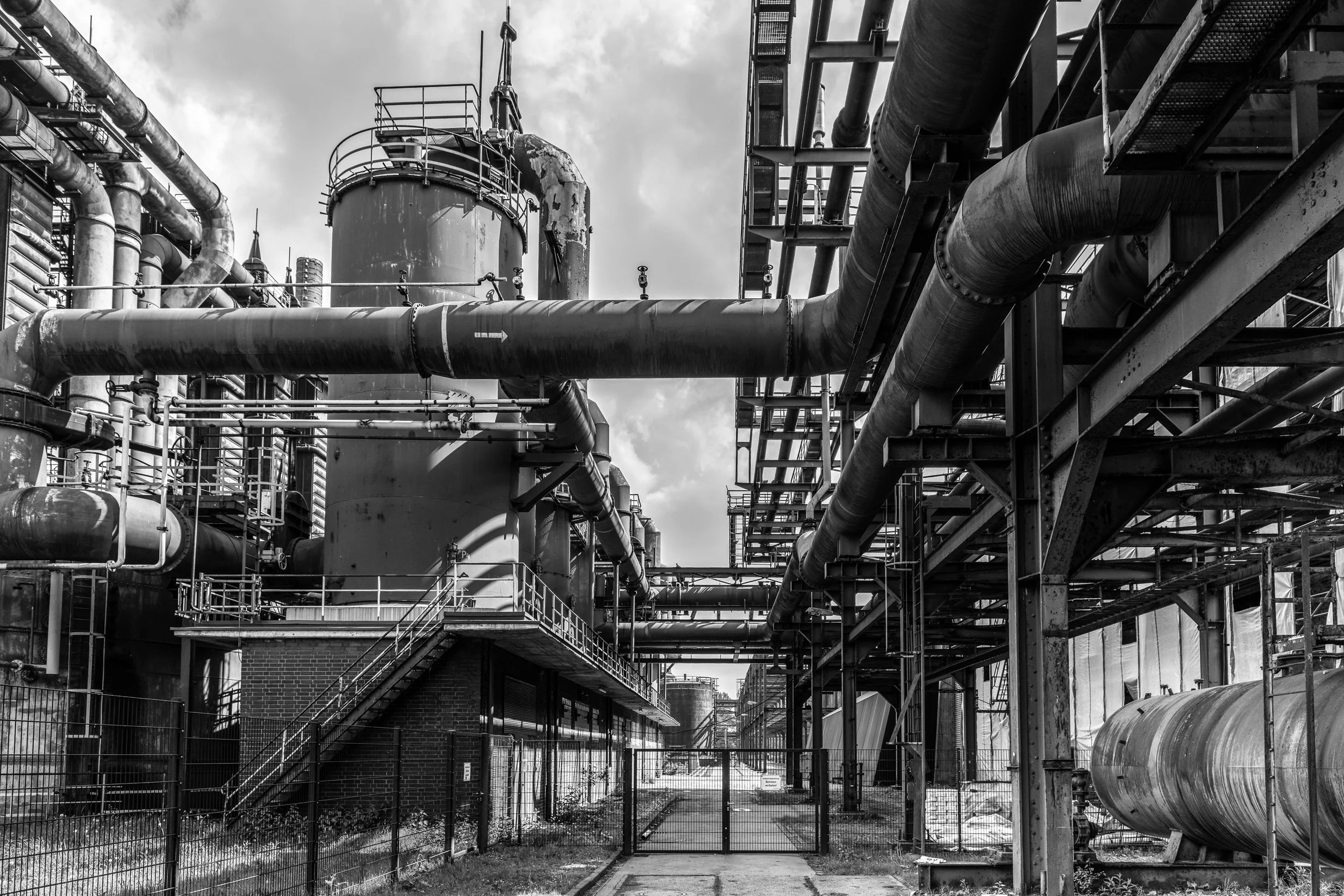 Black and white photo of an industrial factory with large pipes, metal structures, and a fenced gate.