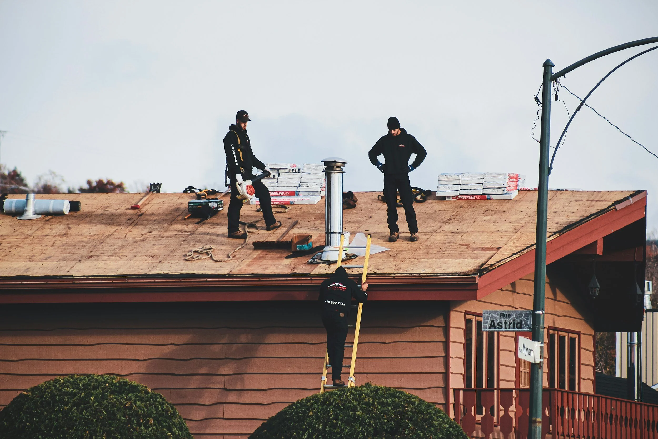 Three workers installing a new roof on a house, with one on a ladder and two on the roof, during daytime.