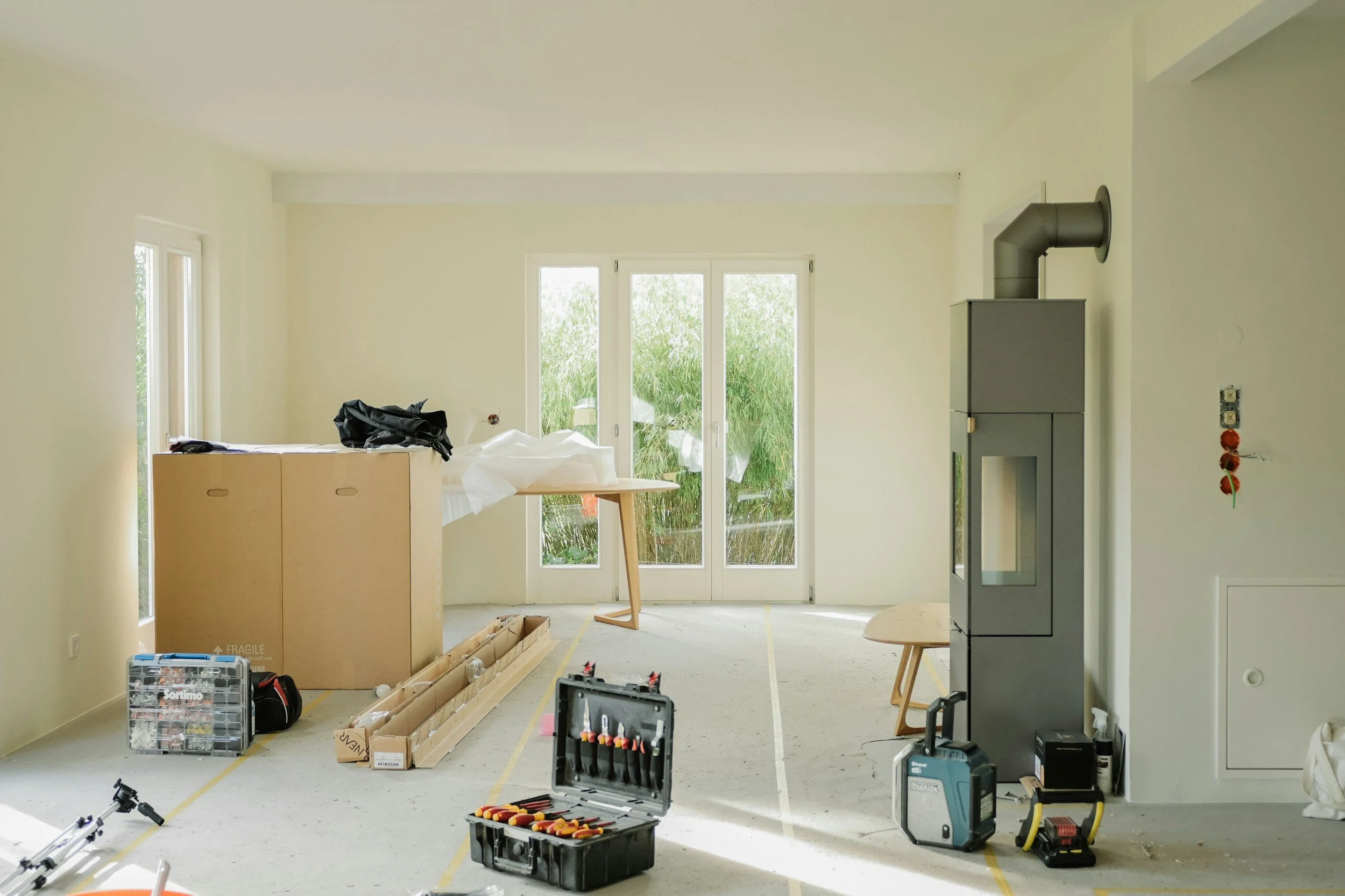 An unfinished living room with construction tools, cabinet, and a wood stove, illuminated by natural light from glass door and windows.