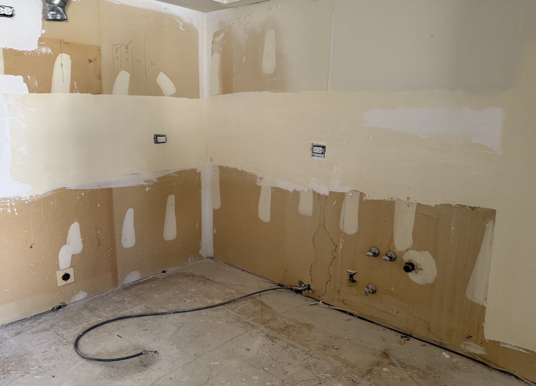 A wall in a kitchen under renovation, with drywall patches, electrical outlets, and plumbing fixtures installed but unfinished, and a black hose on the floor.