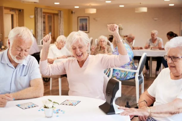 Elderly woman celebrating and smiling with arms raised in a senior center or assisted living facility, surrounded by other elderly residents sitting at tables.