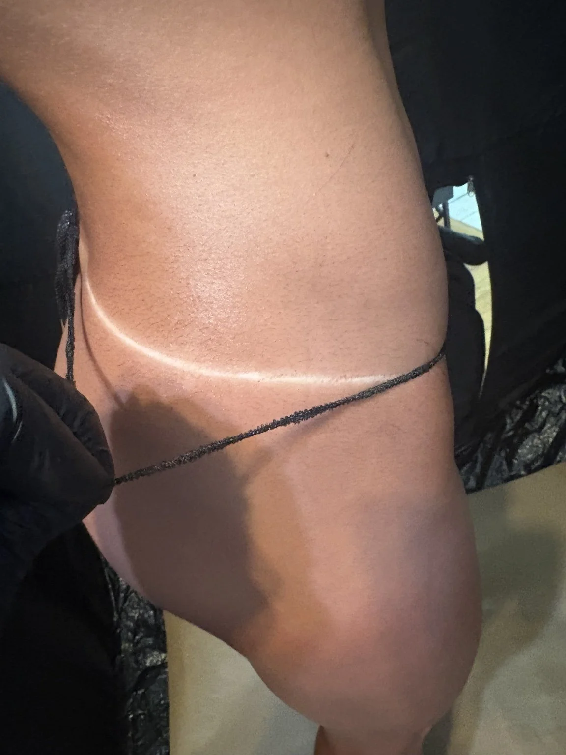 Close-up of a person's lower back and upper thigh, showing a tattoo, and a black chain or string being held in a gloved hand.