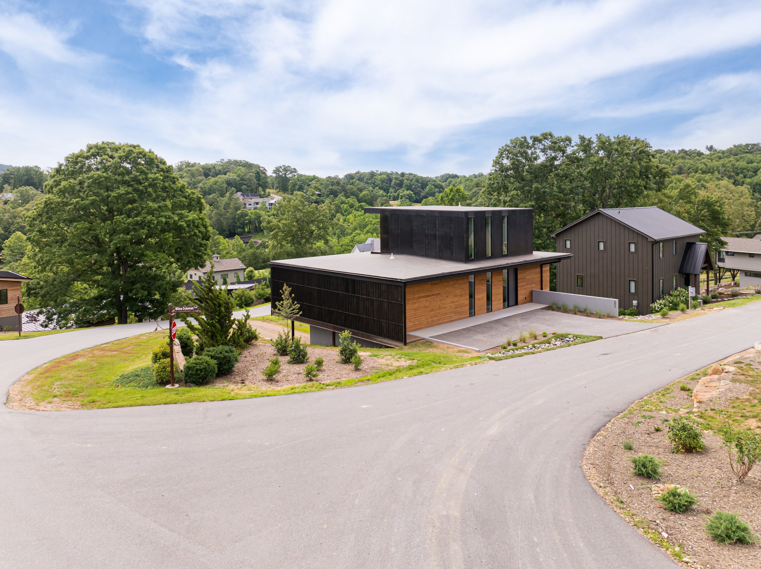 Modern house with black and wood exterior, situated on a curved driveway in a lush hilly neighborhood, with trees and other houses in the background under a blue sky.