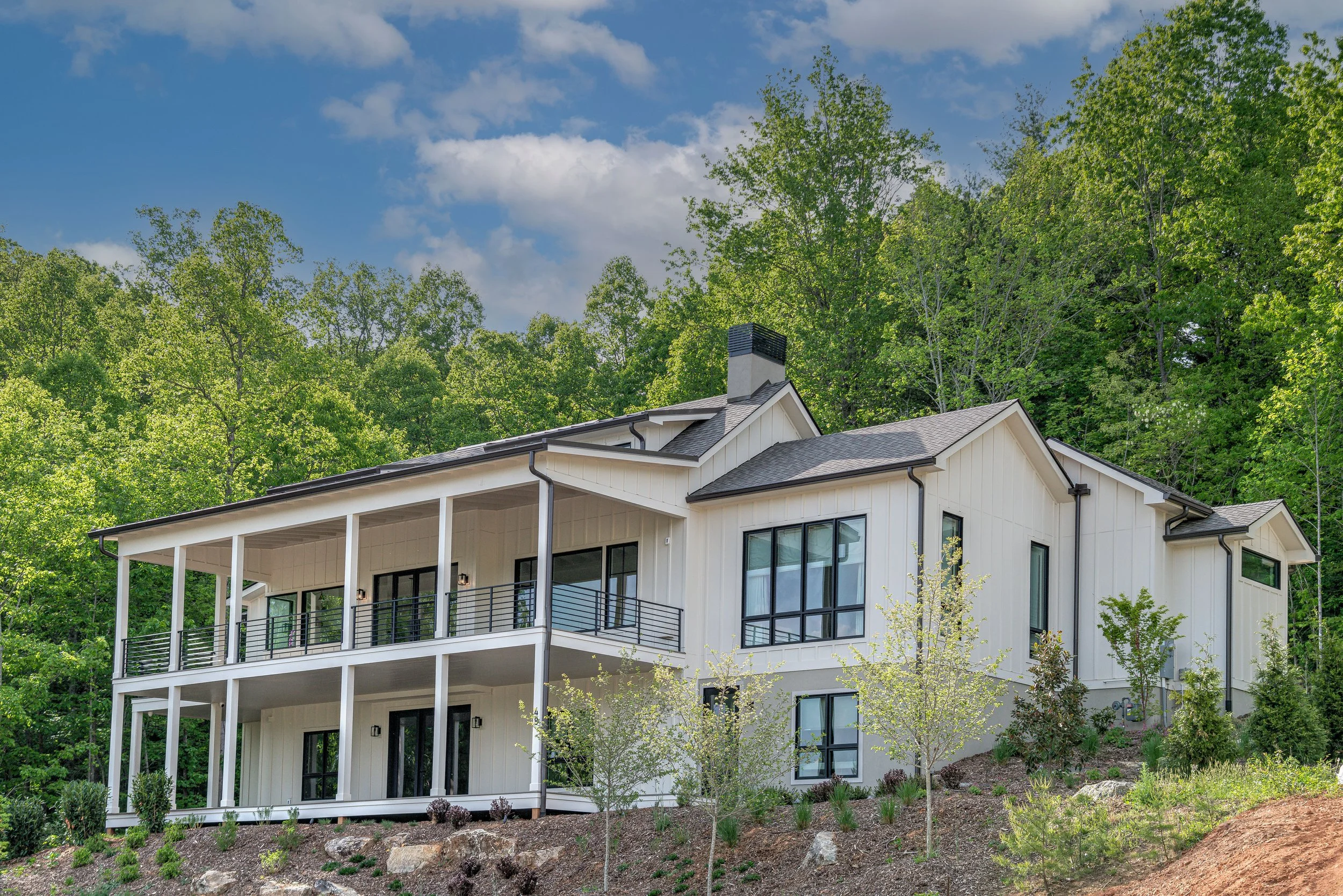 Modern two-story house with large windows and a spacious balcony, situated on a hillside with trees and shrubbery.