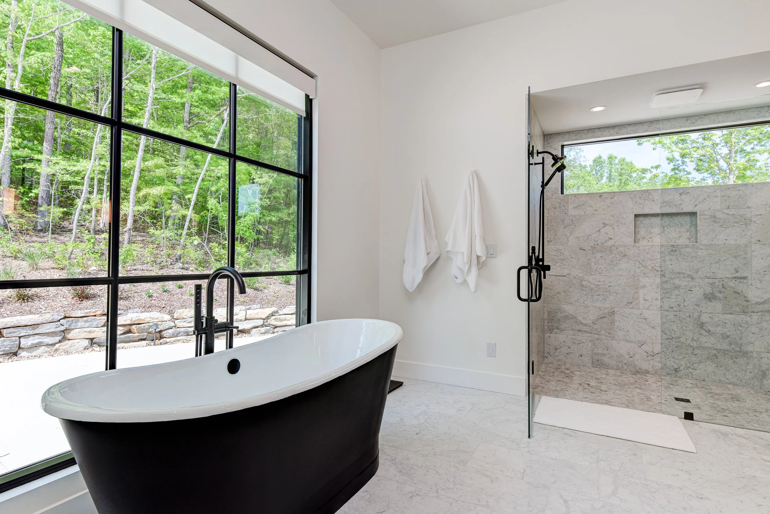 Modern bathroom with a freestanding bathtub near a large window overlooking a wooded area and a glass-enclosed shower with marbled tiles and a window on the wall.