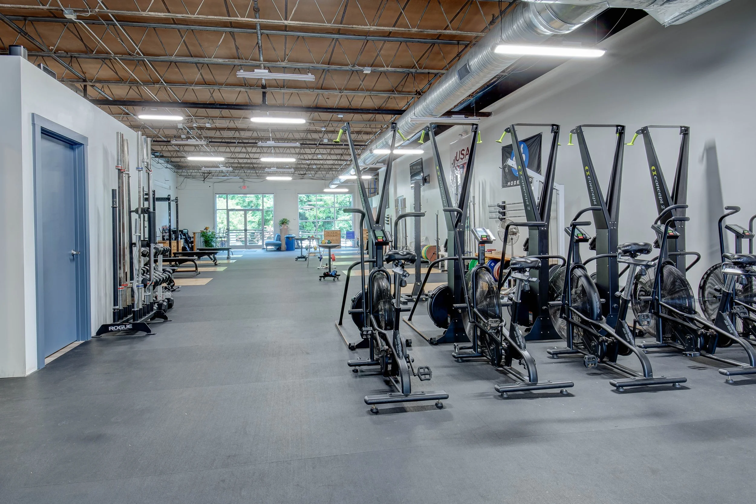 Inside a modern gym with cardio equipment, including ellipticals and other machines, arranged in rows, with large windows at the end letting in natural light, and exposed ceiling pipes.