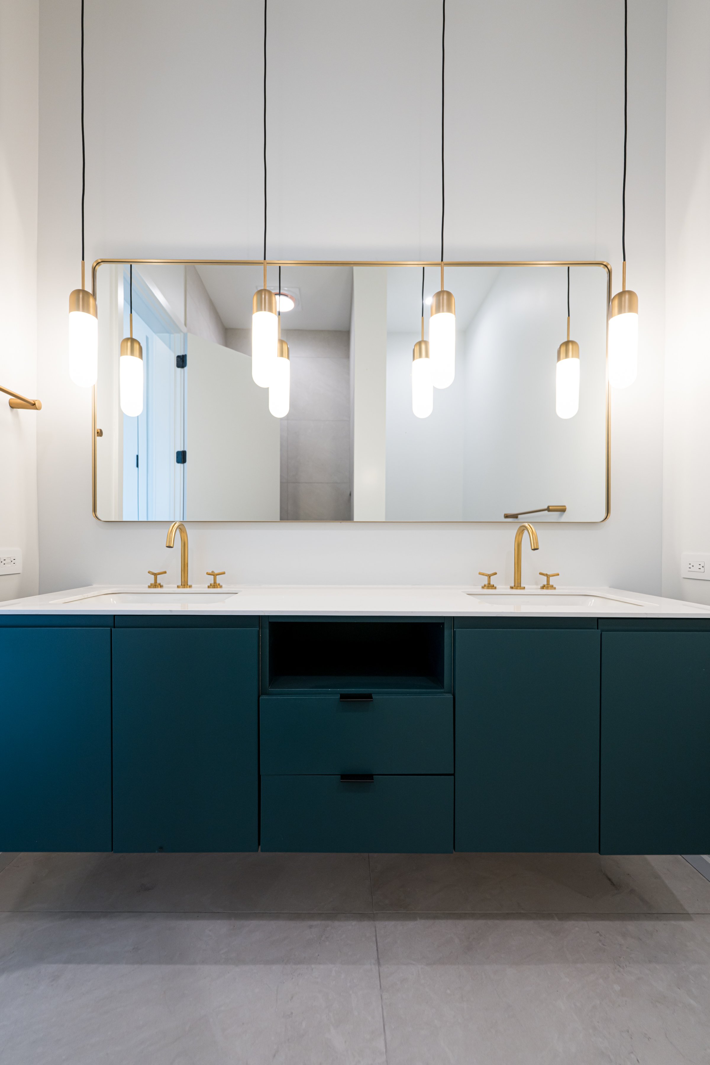 Modern bathroom vanity with doublesinks, gold faucets, teal cabinet, large mirror, and hanging pendant lights.