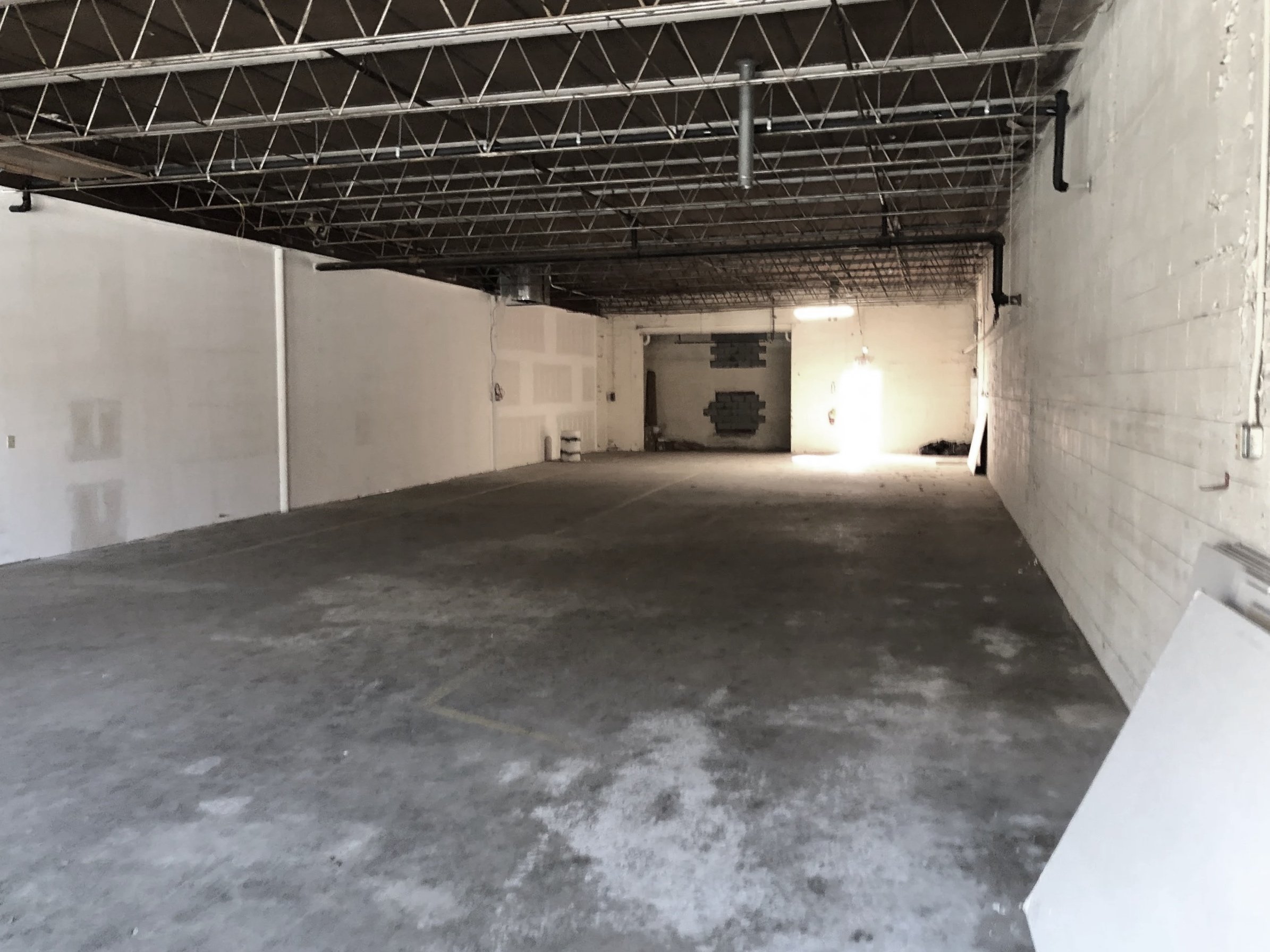 Empty indoor parking garage with concrete floors and white painted brick walls, exposed ceiling beams, and a partially lit back area.