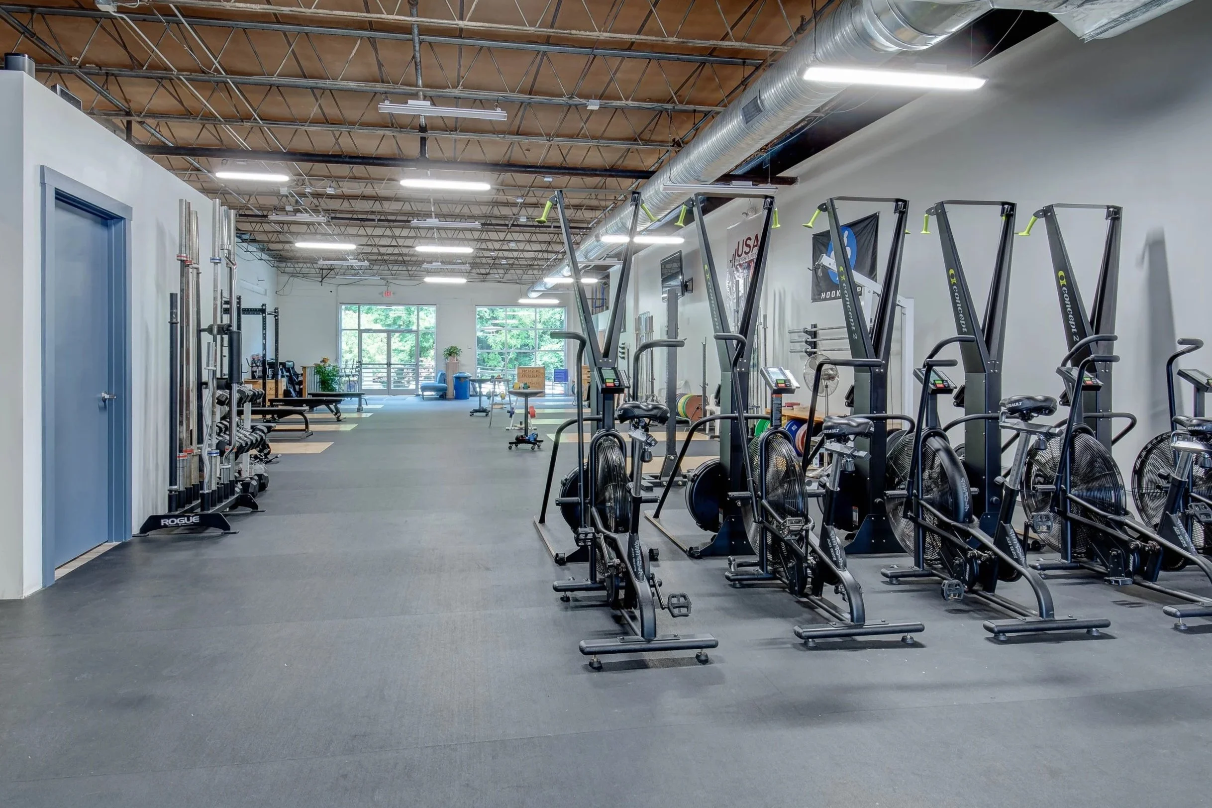 Empty gym with weightlifting equipment, exercise bikes, and sunlight coming through large windows.