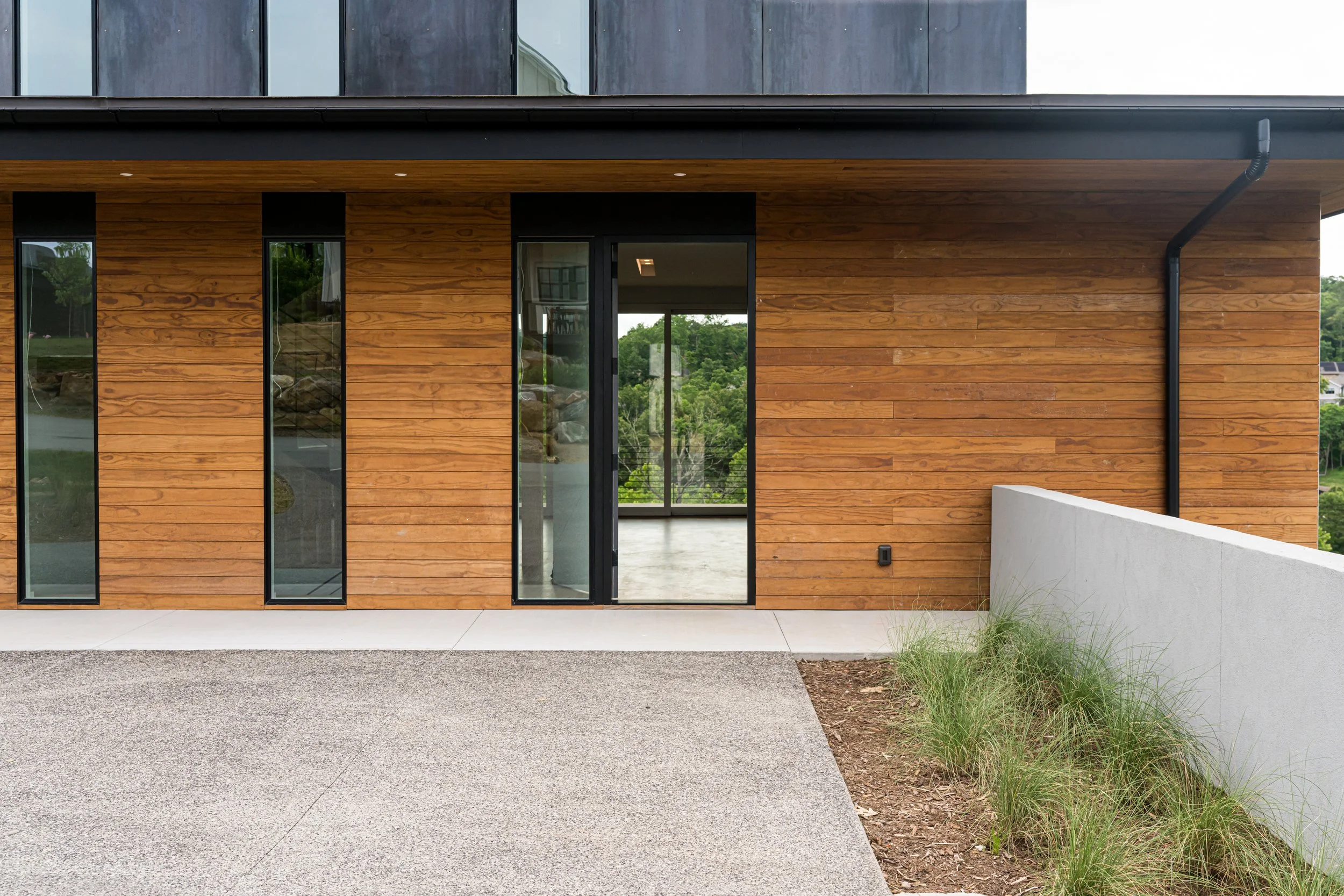 Exterior view of a modern building with a wooden facade, large glass sliding door, and narrow vertical windows, with a concrete patio and some greenery.
