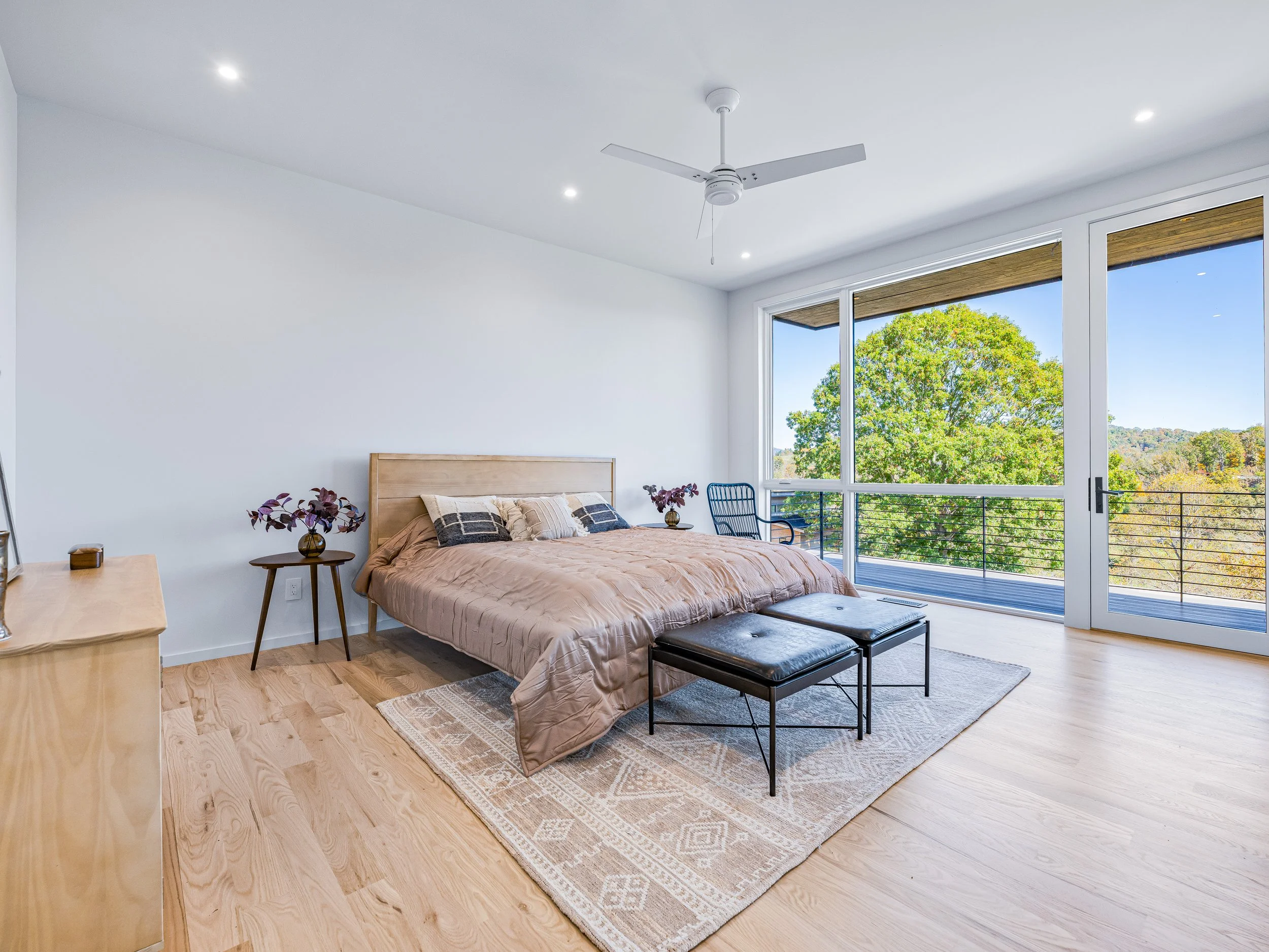 Bright bedroom with a large window showing green trees outside, a bed with a beige comforter and pillows, a nightstand with a vase of flowers, a small chair, two black ottomans at the foot of the bed, a wooden dresser, a ceiling fan, hardwood floorin