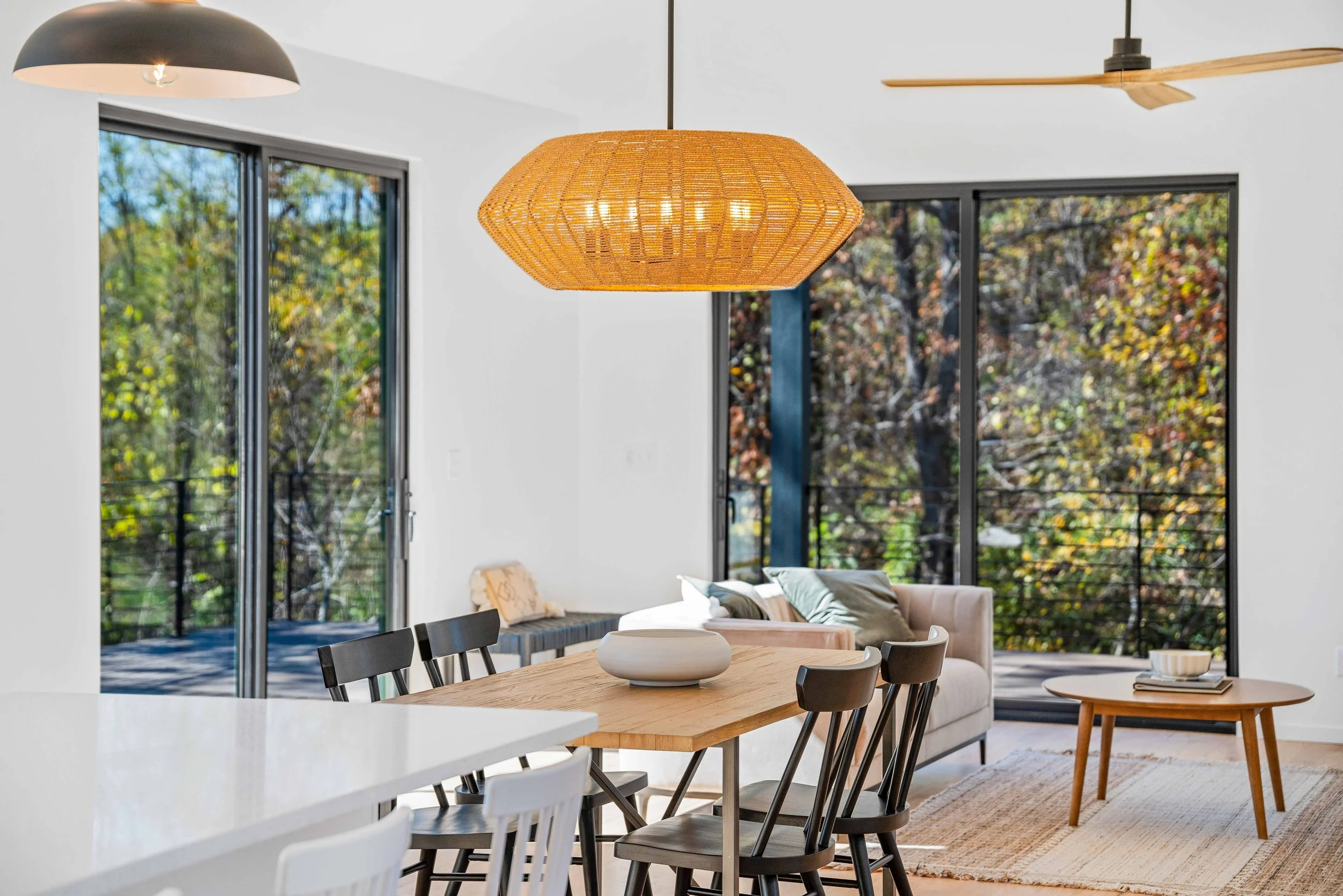 Interior of a modern living room and dining area with large windows showing trees outside, a hanging wicker light, a bookshelf, a sofa with cushions, a wooden coffee table with books, and a ceiling fan.