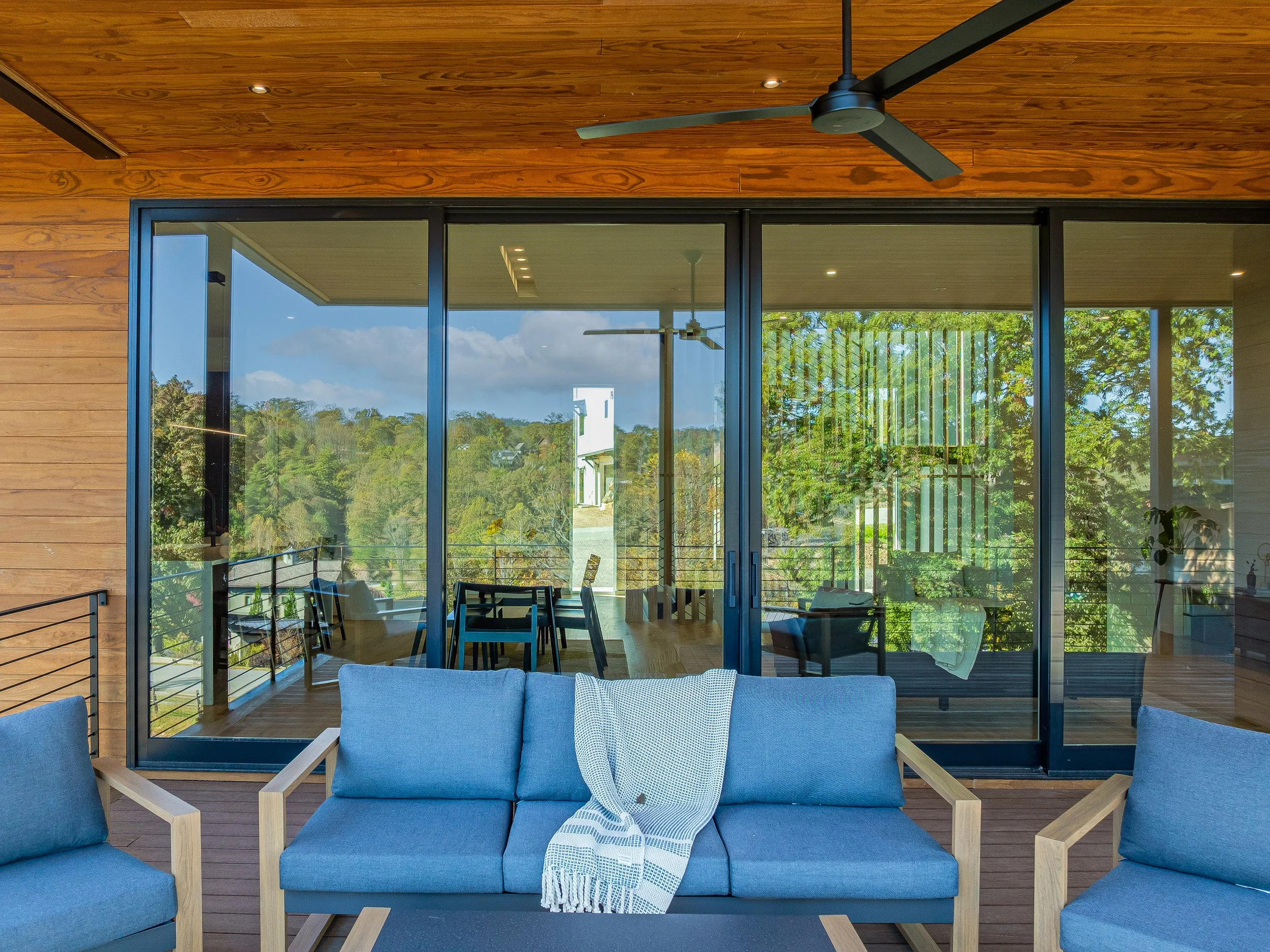 A modern outdoor patio with blue cushioned sofa, wooden armrests, and a throw blanket, overlooking a balcony with glass sliding doors, wooden ceiling, ceiling fan, and lush green trees outside.