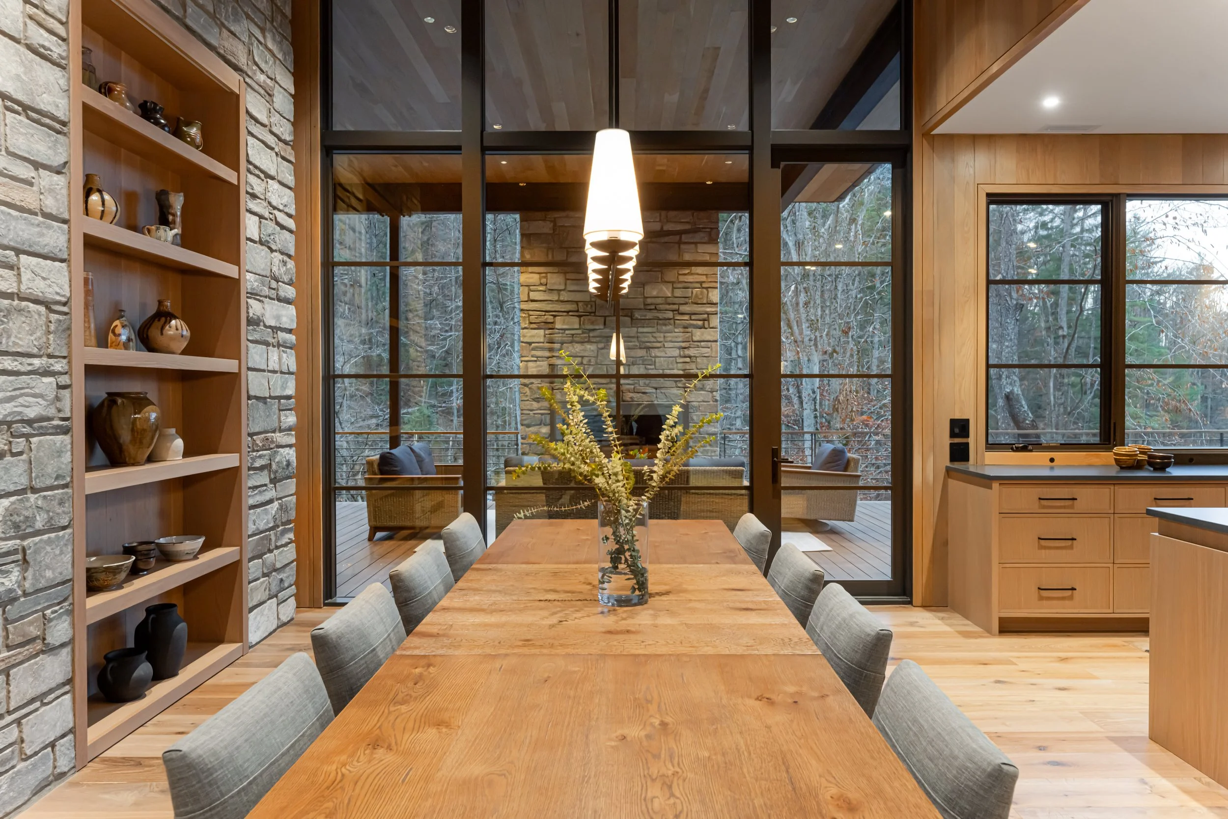 Modern dining room with wooden table, grey chairs, stone and wood walls, large glass windows viewing a wooded outdoor deck with seating, and a pendant light fixture above the table.